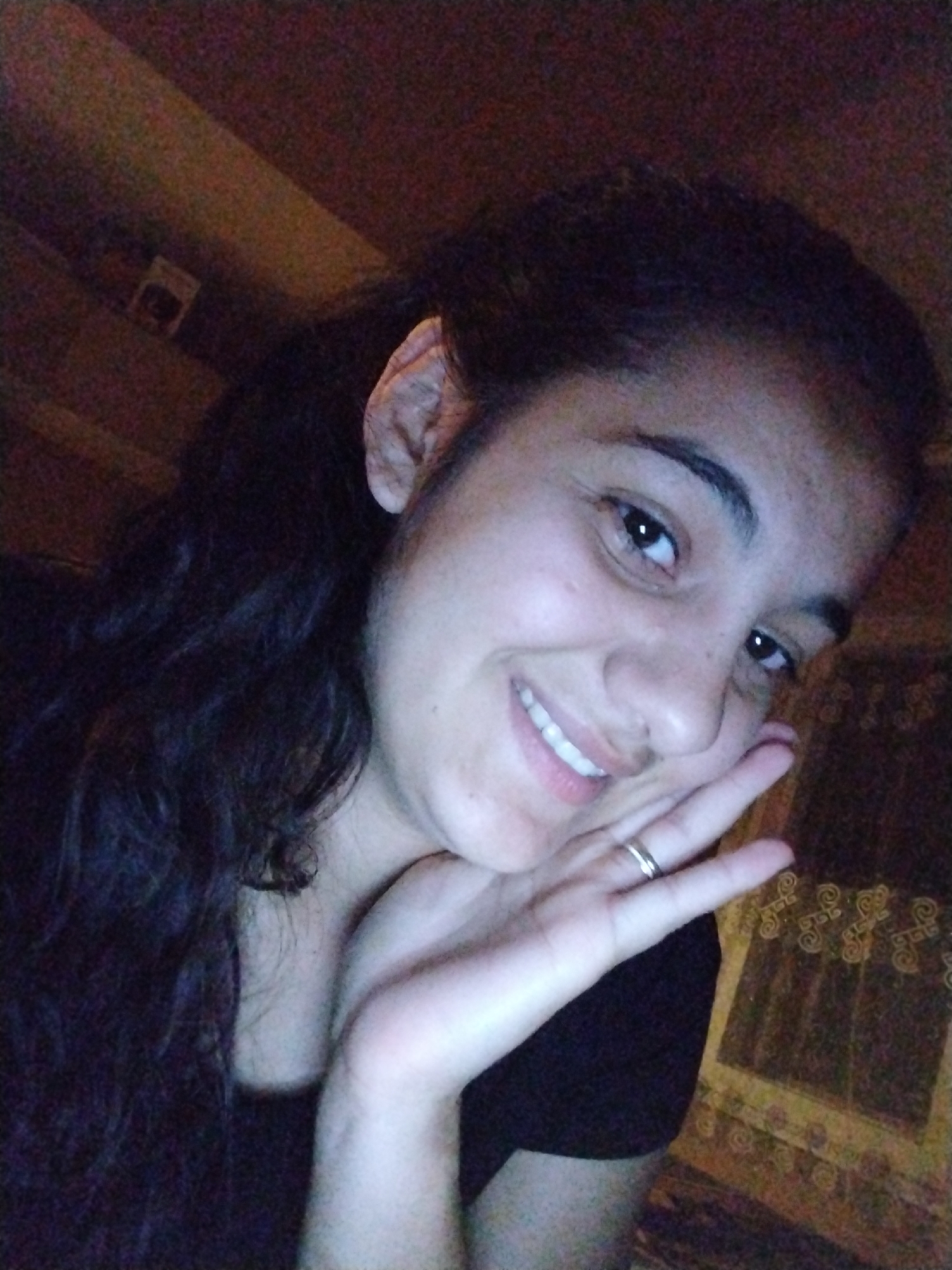 A young woman is prominently featured in a close-up, informal portrait, likely a selfie. She has dark, curly hair, pulled back, and dark eyes. She is smiling broadly, revealing her teeth, and appears to be in good spirits. Her right hand is raised to her face, with her chin resting on her palm, and she wears a silver-colored ring on her ring finger. Her skin tone is medium, and she has visible pores and minor skin texture. She is wearing a dark-colored top.
The setting appears to be indoors, with the background being mostly dark and out of focus. Hints of warm, possibly wooden or fabric textures, and faint patterns are visible behind her, suggesting a home or casual indoor environment. A small, indistinct rectangular object can be seen in the upper left background. The lighting is soft and somewhat dim, casting warm tones on her face, indicating an indoor setting with artificial light or low natural light. There is no visible text. The overall mood of the image is friendly and relaxed, conveying happiness through her smile.