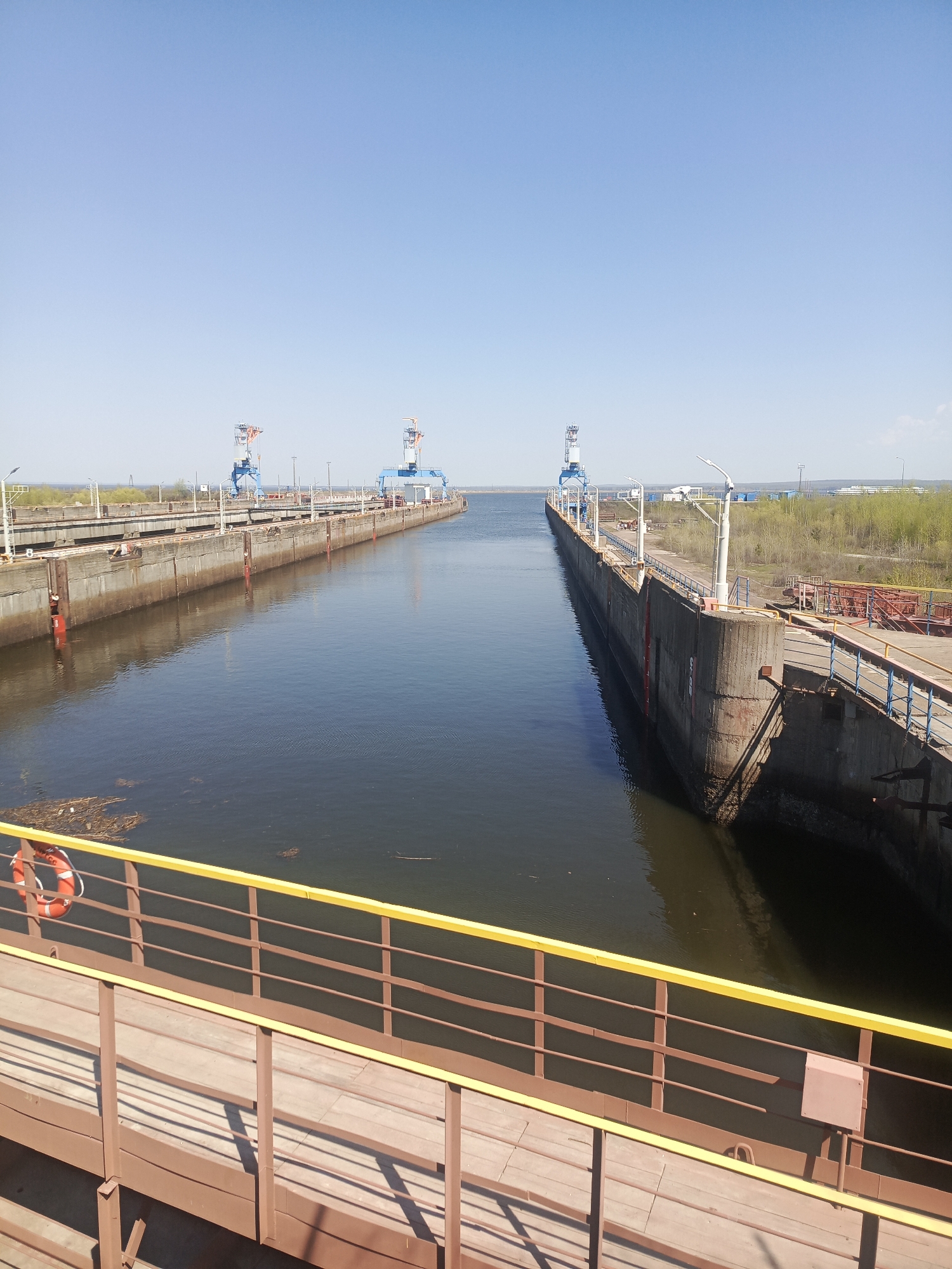 This image shows a waterway in Chaikovsky, Russia. The waterway has two concrete locks with metal gates, and the locks have blue and red cranes. The water is dark and still. The sky is blue and clear. There is a wooden walkway with a yellow railing in the foreground. There is a life preserver hanging from the railing. In the background, there are trees and buildings.