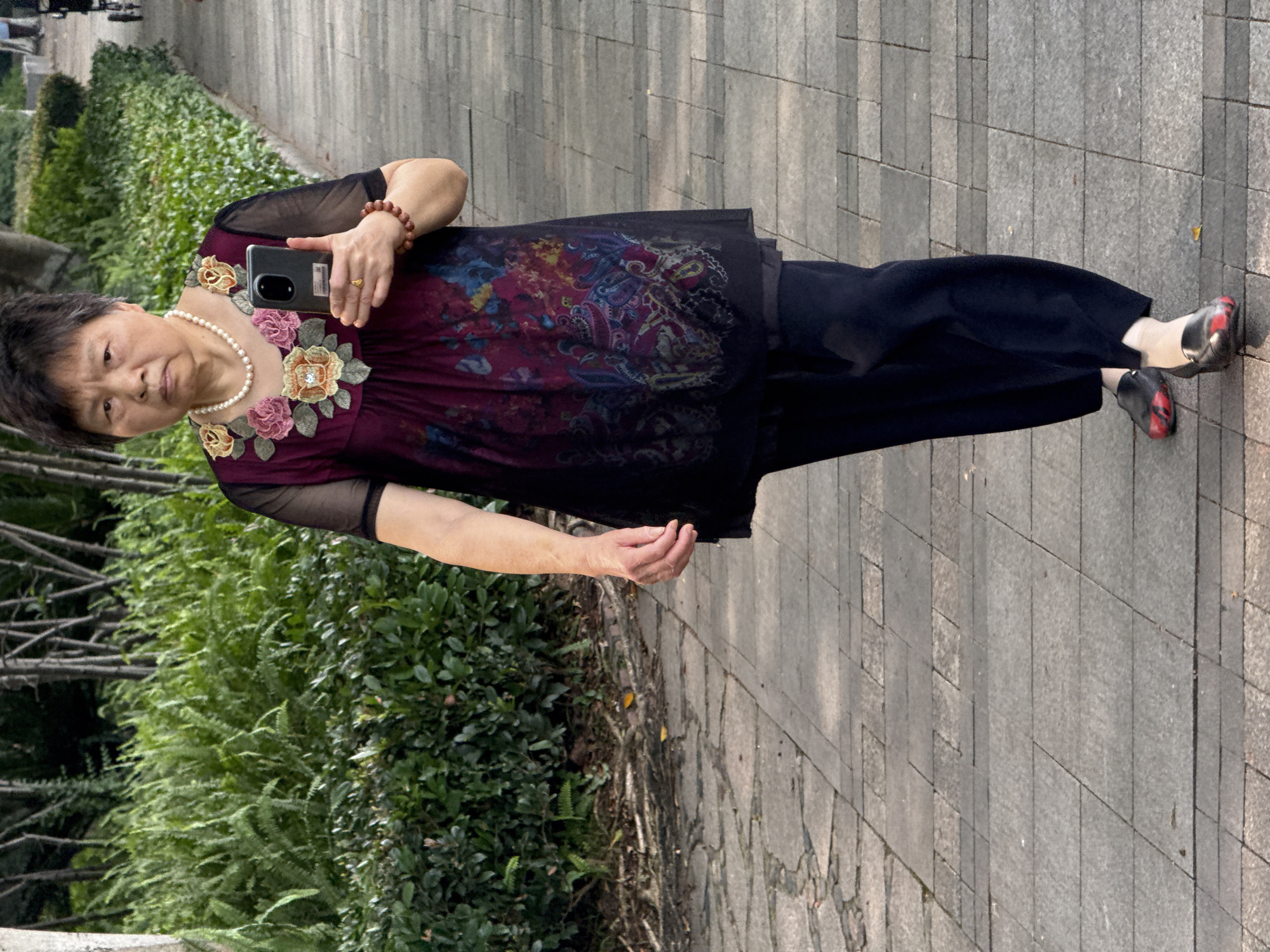 An Asian woman, appearing to be elderly, walks forward on a paved pathway in an outdoor park in Jiangjin, China. She has short, dark grey hair and a neutral expression as she looks directly ahead. She wears a pearl necklace and a dark, sheer top with a patterned underlayer. The top features elaborate floral embroidery around the neckline in shades of pink, yellow, and green, some with central embellishments, and a colorful, abstract pattern in blues, yellows, and reds on its body. She wears dark trousers and dark loafers with prominent red accents on the toes. Her right wrist bears a reddish-brown beaded bracelet and she holds a dark-colored smartphone vertically in her right hand, with the screen facing away. A gold-toned ring is visible on her right ring finger. The pathway consists of light grey rectangular paving stones. To the left, dense green bushes and ferns line the path, with bare tree trunks visible further in the background. To the right, a similar line of manicured green bushes borders the path, and a blurred wheeled object, possibly a stroller or wheelchair, is faintly visible in the distant background.