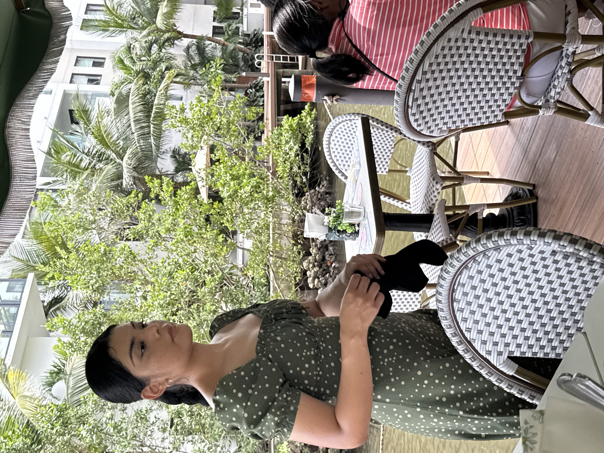 The image captures a scene at a waterfront restaurant in Fort Lauderdale, Florida, United States of America. A woman wearing a green dress with white polka dots is seated at a table. She is holding a black object, possibly a wallet or clutch. Nearby, two other people are seated at a table, one wearing a red and white striped shirt. The restaurant features wicker chairs. The backdrop shows lush greenery, palm trees, and buildings, with a waterway running adjacent to the dining area.