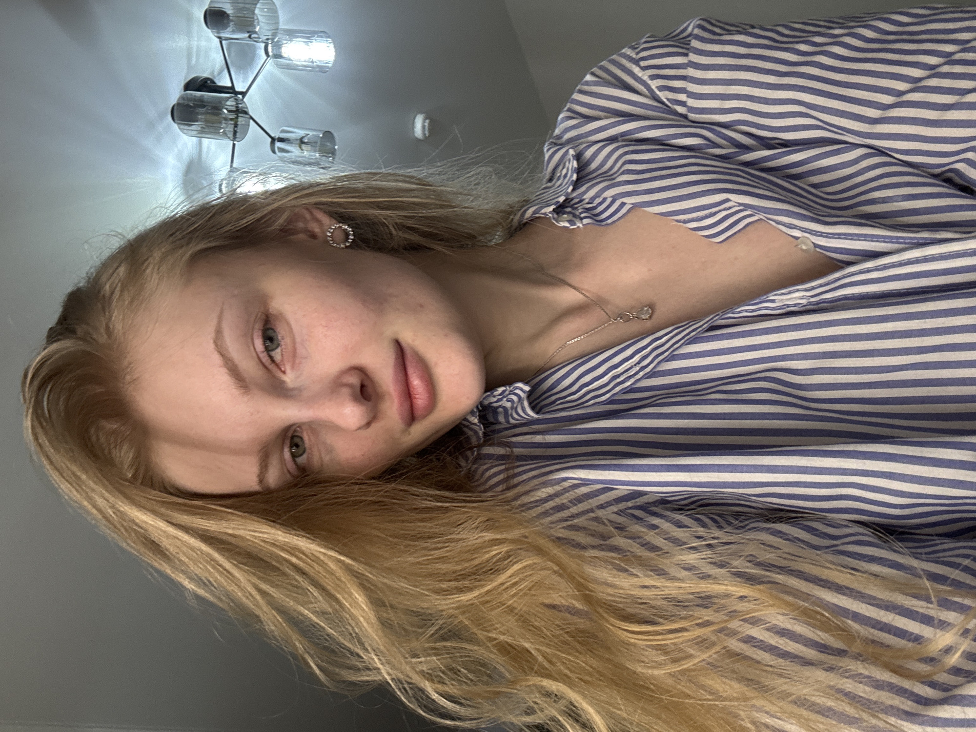 A fair-skinned young woman with long blonde hair is angled towards the viewer, wearing a blue and white striped shirt, a round earring in her left ear, and a delicate silver necklace. The background consists of a light gray wall with a modern chandelier. The city of Perm, Russia, is not visually discernible in the image.