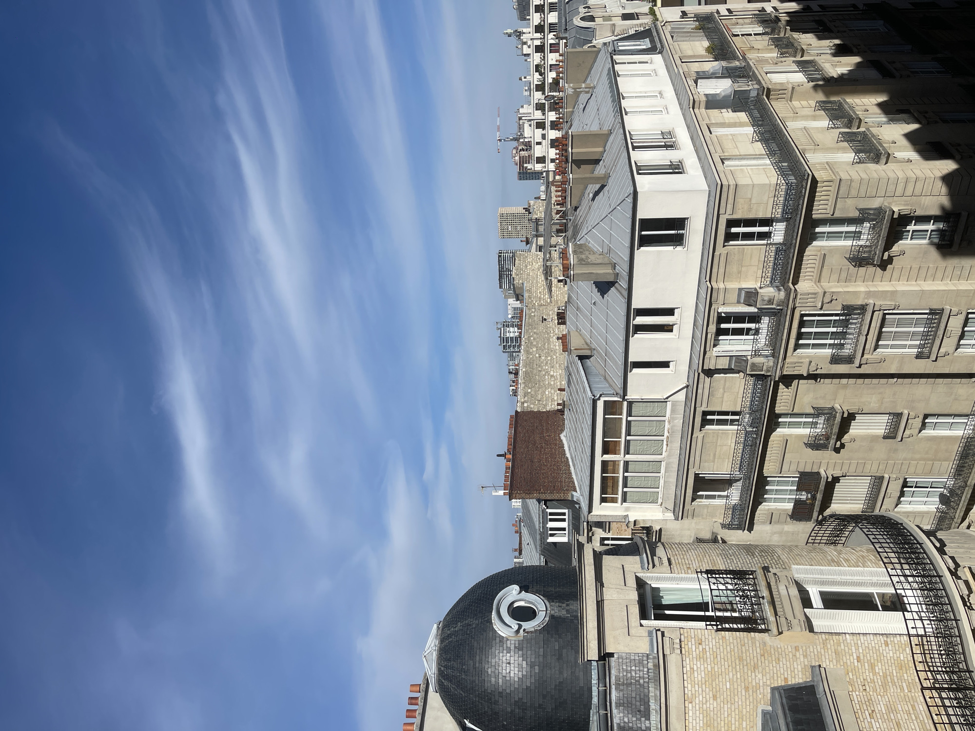 The image captures a rooftop view of Parisian architecture under a bright blue sky with wispy white clouds. The foreground is dominated by the mansard roofs and ornate facades of classic Haussmannian buildings, characterized by their stone exteriors, numerous windows, and wrought-iron balconies. A distinctive dark, domed structure with a circular window is prominently positioned on the left, adding a unique architectural element to the scene. In the mid-ground and background, a cityscape unfolds with more residential buildings, some appearing to be under construction with cranes visible. The overall impression is of a sunny day in an urban environment, likely Paris, given the architectural style. There are no people visible in the image.