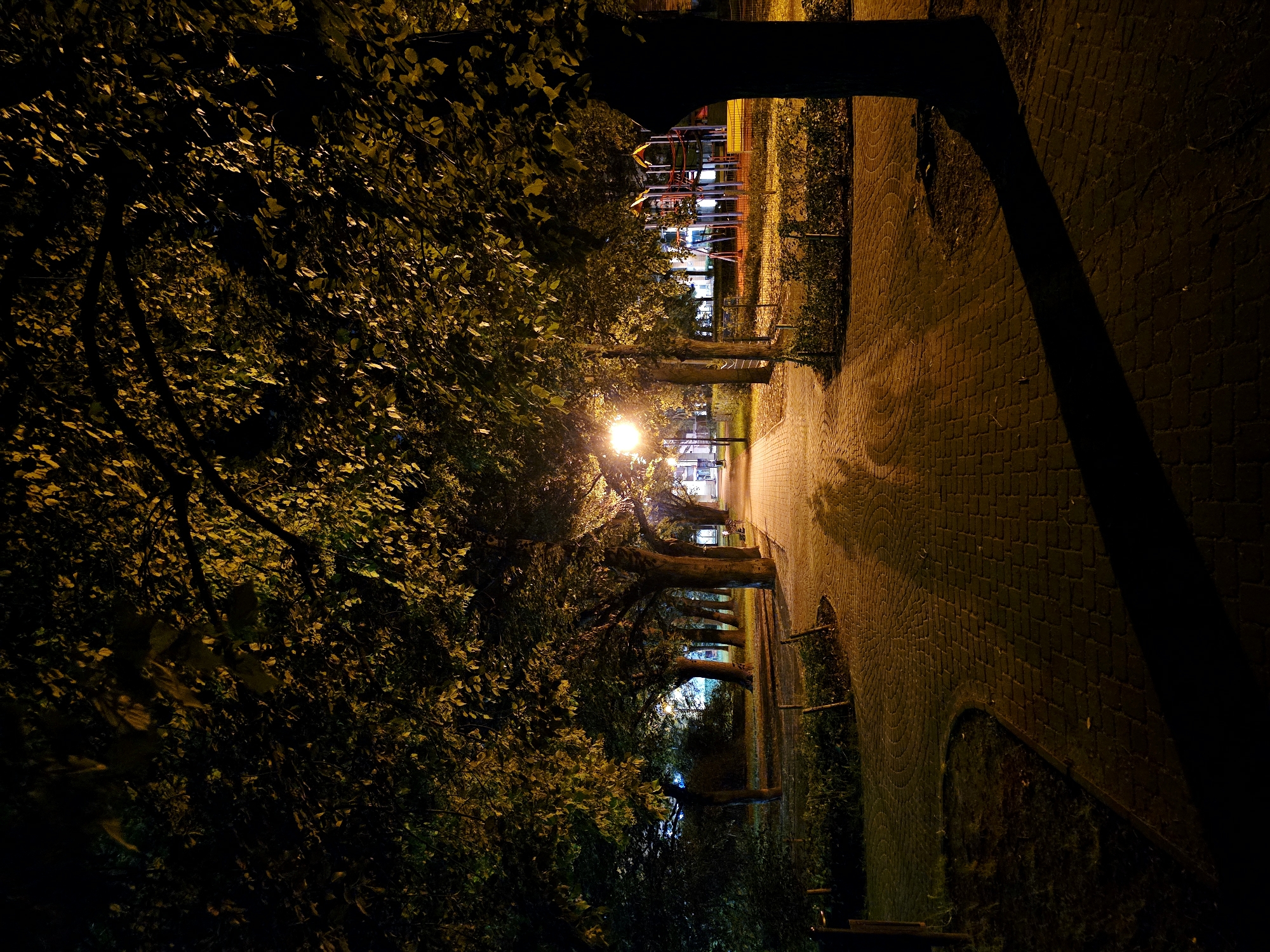An illuminated pathway in a park at night, likely in Warsaw, Poland, is the subject of this image. The path, paved with interlocking bricks, curves gently into the distance, bordered by trees and grassy areas. A streetlamp casts a warm glow on the path, creating long shadows. In the background, a playground with brightly colored equipment is visible, hinting at a community space. The overall atmosphere is serene and quiet, suggesting the park is empty at this hour. The presence of a playground indicates a family-friendly location.