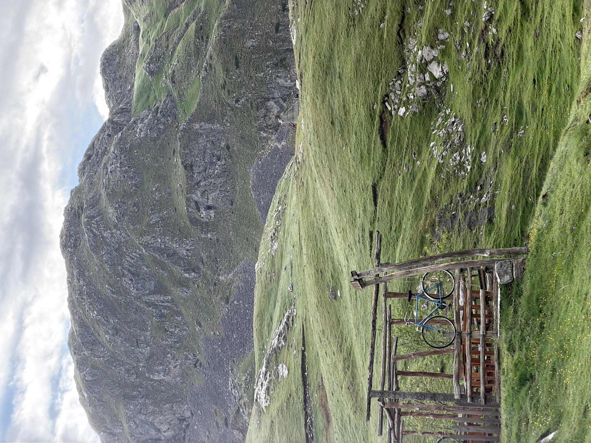 The image captures a rugged, mountainous landscape, characteristic of Kapetanovo Jezero, Montenegro. In the foreground and slightly to the right, a rustic wooden structure, possibly a shelter or an observation point, is prominently featured. A bright blue bicycle rests within this wooden framework. The ground around the structure is covered in lush green grass, dotted with small yellow wildflowers.
Beyond this immediate area, the terrain slopes upwards and transitions into steep, imposing mountains. These mountains are predominantly grey and rocky, with significant patches of vibrant green grass and vegetation clinging to their slopes and crevices. The texture of the rock faces is detailed, showing weathering and geological formations. In the middle distance, a narrow path or track is discernible winding through the mountainous terrain.
The sky above is overcast with soft, diffuse clouds, suggesting a mild, possibly cloudy day rather than bright sunshine. No people are visible, lending a sense of peaceful solitude to the scene. The image evokes a feeling of natural grandeur and outdoor activity. There is no visible text.