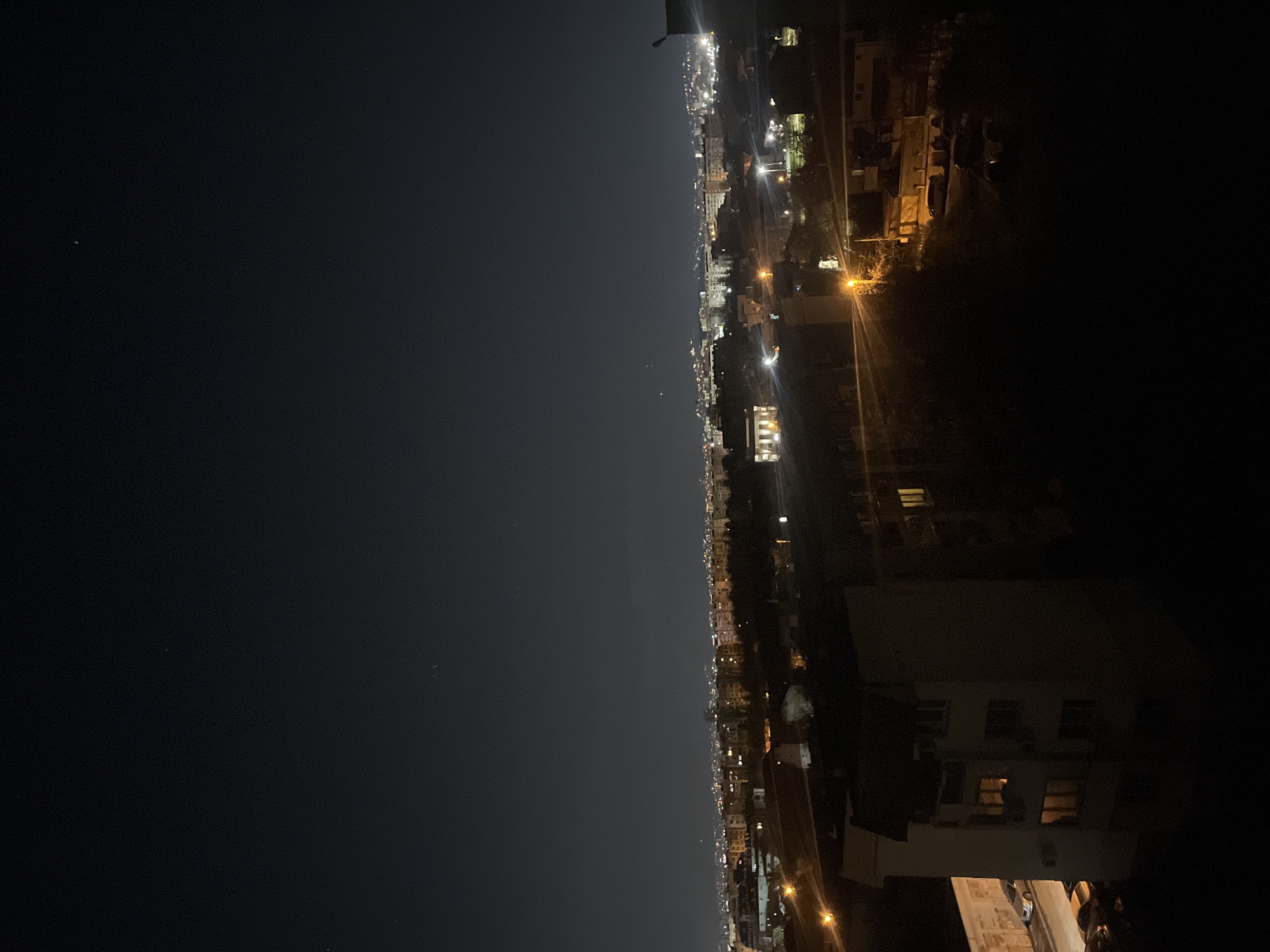 The image captures a nighttime view of a city from a high vantage point, possibly Sofia, Bulgaria, as implied by the presence of urban structures and lighting consistent with a city environment. The left side of the image displays a dark sky, which transitions into a cityscape lit by streetlights and building illumination on the right. Buildings of varying heights are visible, densely packed together and extending into the distance. There is no clear indication of human activity in the image. The overall environment appears to be an urban residential area at night.