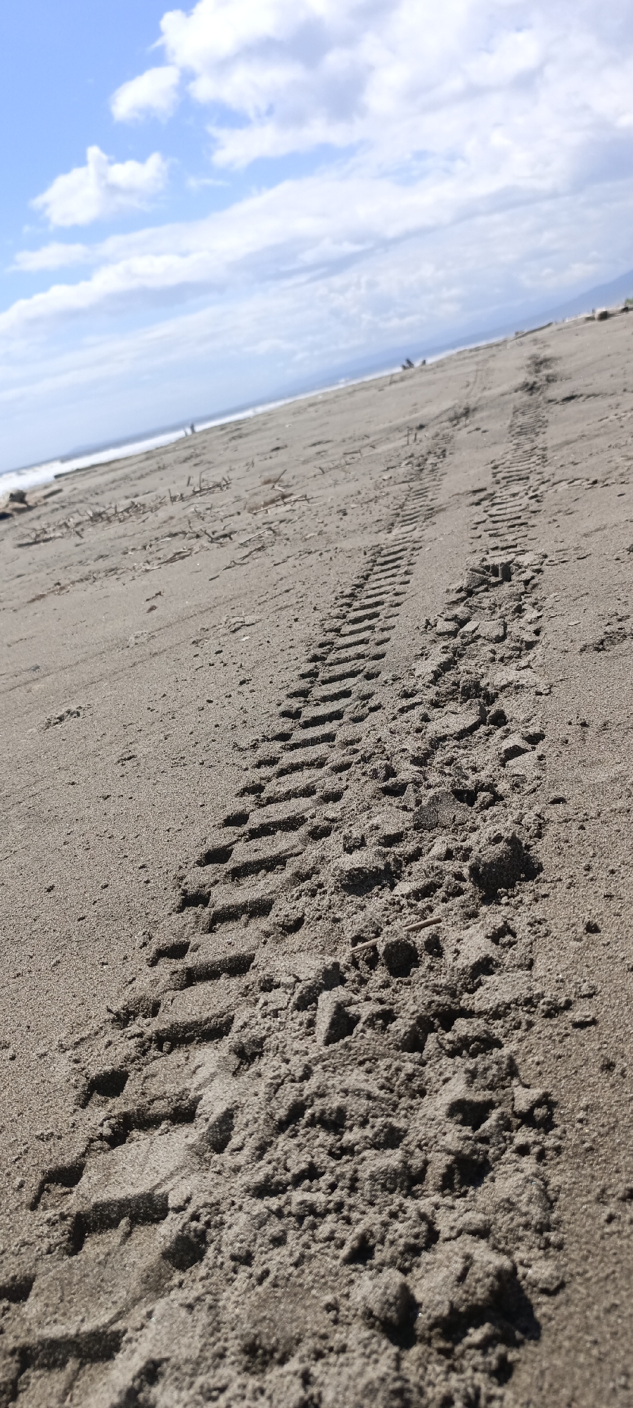 An outdoor, close-up, vertical, low-angle shot shows tire tracks in a sandy beach under a partly cloudy sky. The tire tracks are prominent in the foreground and are running diagonally from the bottom right to the upper left. The sand is light brown and textured with small indentations and some small pieces of dried vegetation. In the distance, the ocean meets the horizon, with a line of white surf. The sky above is blue with scattered white clouds. There are no people clearly visible in the frame.