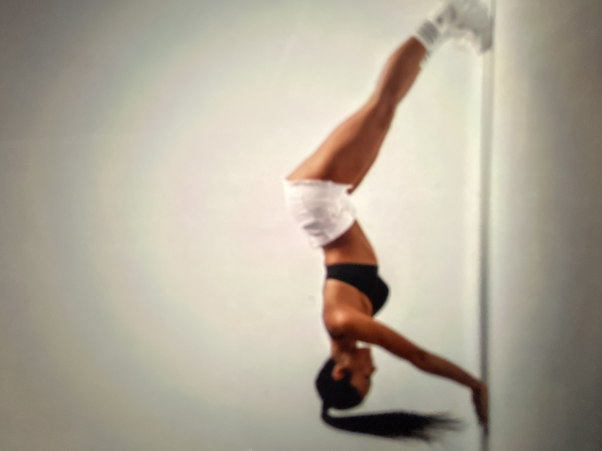 The media file shows a person performing a handstand against a wall. The subject is a woman with dark hair, wearing a black sports bra and white shorts, and white socks and shoes. Her body is inverted, with her hands on the floor and her legs extended upwards, touching the wall. Her arms are straight, and her body forms a long, diagonal line. The background is a plain, light-colored wall, suggesting an indoor setting like a gym or studio. The lighting is soft and even, with no harsh shadows, indicating it's likely artificial light. There is no visible text in the image. The overall impression is one of strength, balance, and athletic prowess. The image does not provide any specific cues to suggest a particular time of day, weather, or precise location, beyond the general context of an indoor space suitable for exercise.