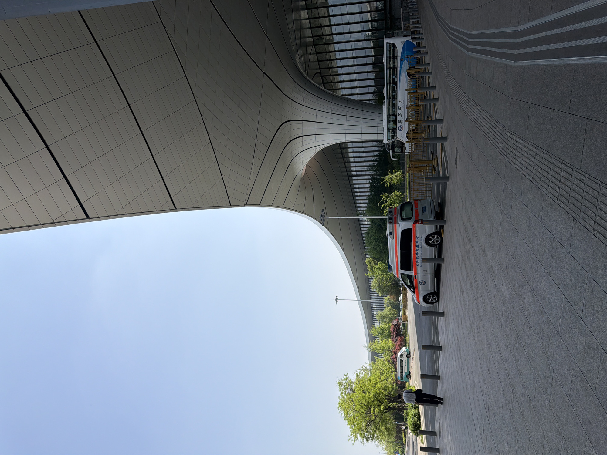 The image shows a view under a curved, modern architectural structure, likely part of a building. A vehicle resembling an ambulance is parked in the foreground. Further back, a larger bus is visible. A person is walking away from the viewer on the sidewalk. The sky is clear, and trees line the street. The location is likely in Langfang, China.