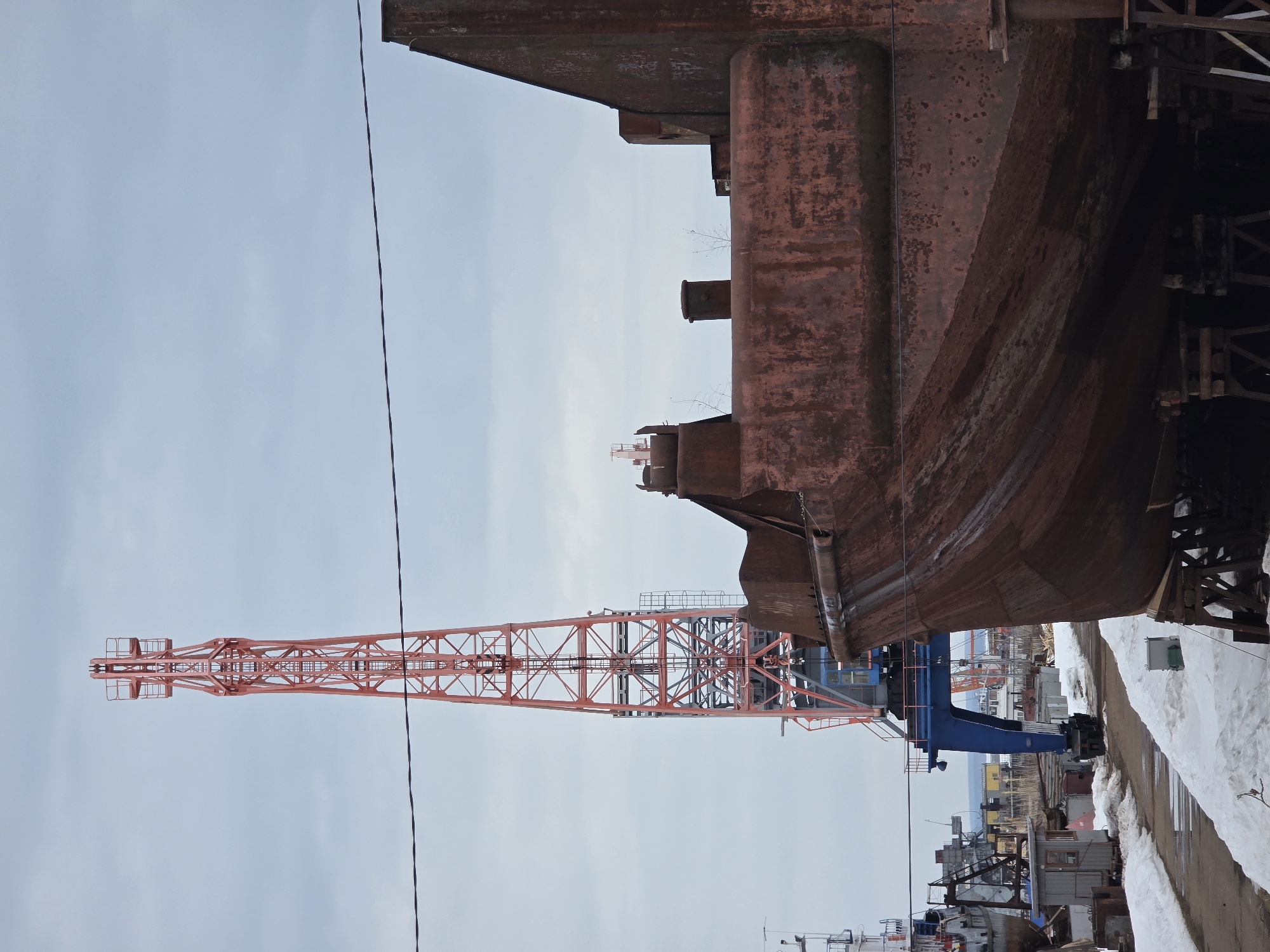 The image shows a shipyard in what could be a cold environment, possibly Chaikovsky, Russia. The main subjects are a large, rusty hull section of a ship under construction or repair, and an orange lattice boom crane extending into a cloudy sky. The ship hull appears to be supported by a blue crane-like support structure. Snow is present along a road or walkway. In the background, there are structures and other elements typical of an industrial shipyard. There's no clear sign of human activity or people in the immediate view.