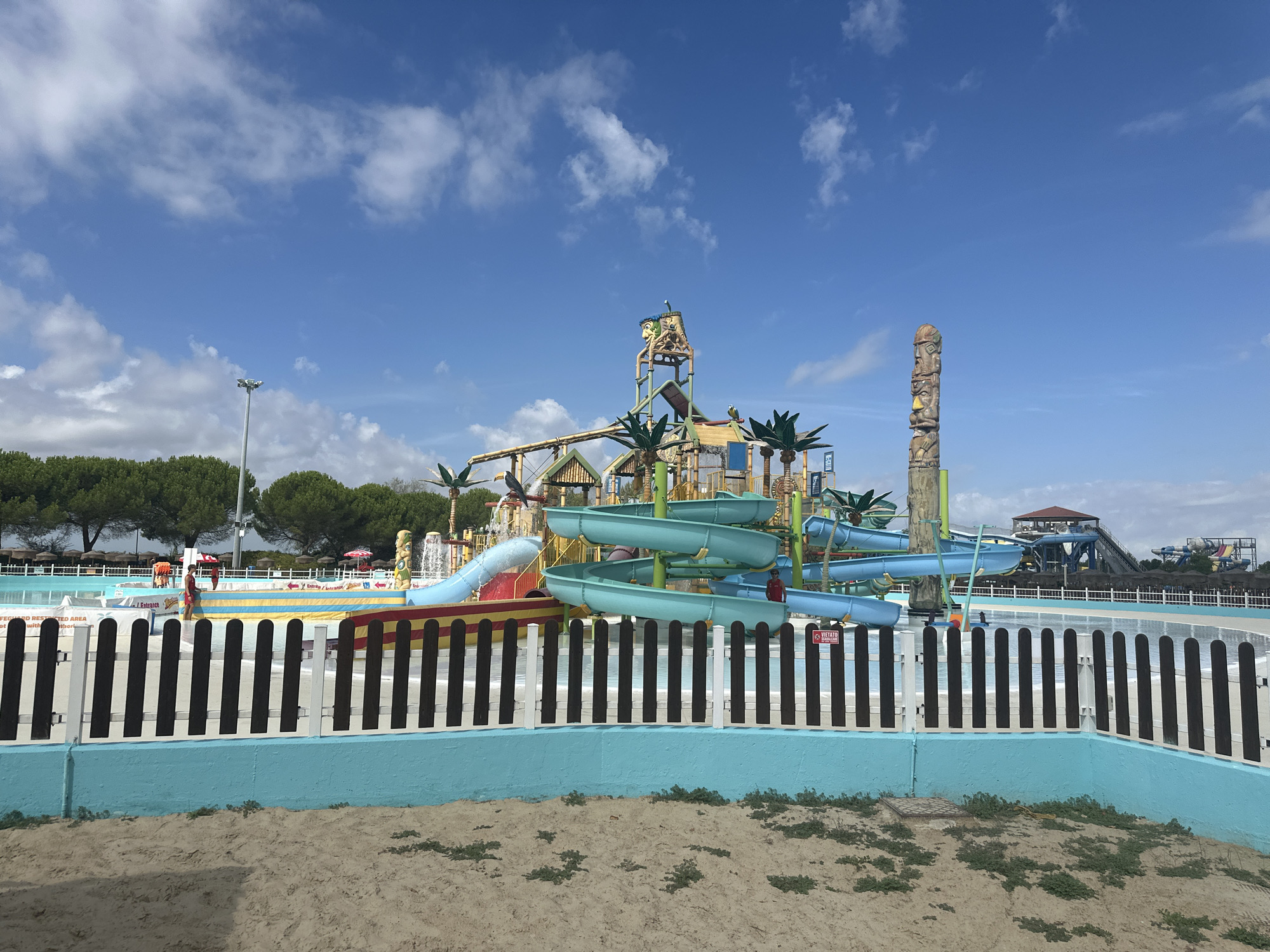 The image shows a water park in Ravenna, Italy. In the foreground, a light blue wall borders a black and white wooden fence, with sand and sparse vegetation in front. Behind the fence, a water park is visible with blue and green water slides, palm trees, and a totum pole. In the background, there are more water slides and trees. The sky is blue with scattered clouds. There are no people clearly visible.