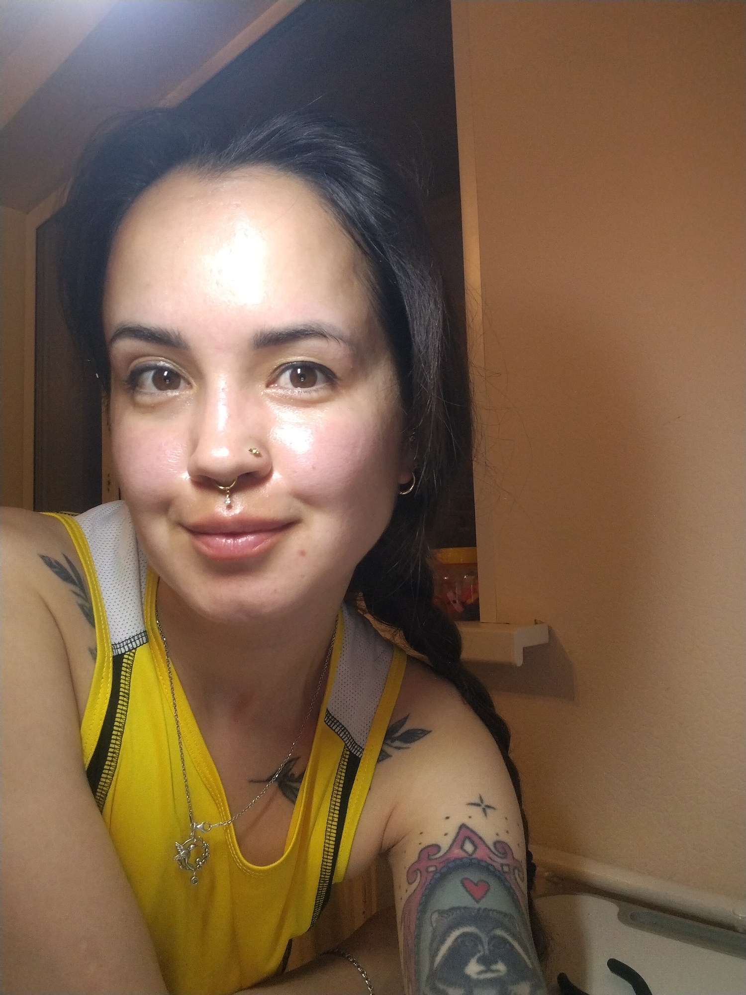 A person with dark hair, brown eyes, and multiple facial piercings (nose and lip) is looking at the camera and smiling slightly. They are wearing a yellow tank top with white trim, and a silver necklace with a charm. The person has tattoos on their arm, one of which depicts a raccoon within a decorative frame, and another of a pair of leaves. The background includes a doorway and a plain wall. A shelf is visible with small containers on it. The image provides no verifiable information that ties the photo to Noviy, Russia.