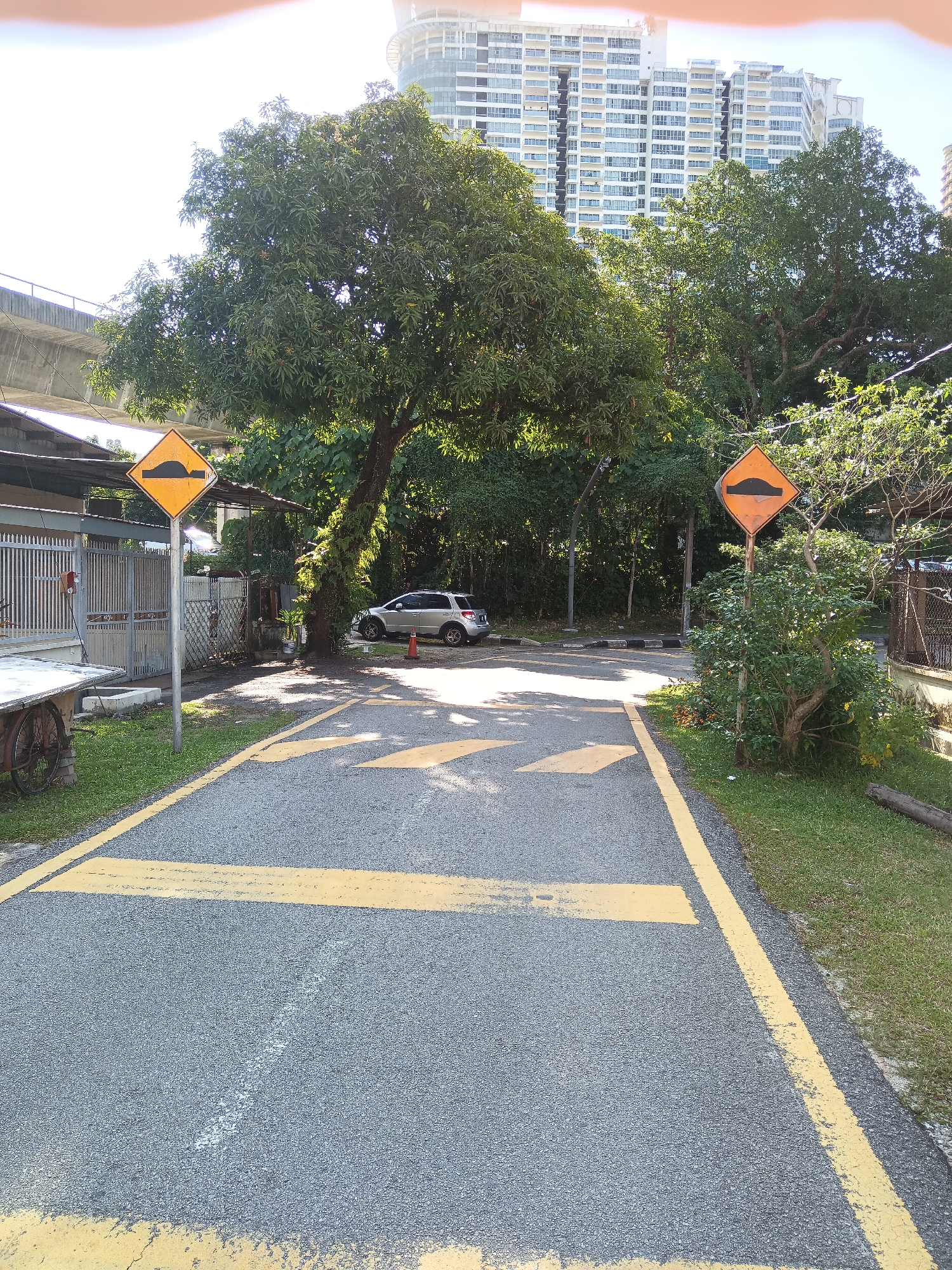 The image depicts an outdoor scene on a paved road with yellow markings, likely in a suburban or transitional area of Kuala Lumpur, Malaysia. A silver hatchback car is parked on the right side of the road, accompanied by a traffic cone. Two diamond-shaped yellow warning signs, each depicting a speed bump symbol, are prominently placed on either side of the road. The road itself is bordered by green grass and trees, with a glimpse of a tall, modern apartment building in the background, suggesting an urban setting. A metal fence and a covered structure, possibly a stall or shelter, are visible to the left of the road. The overall impression is one of a quiet, sunlit day with clear visibility.