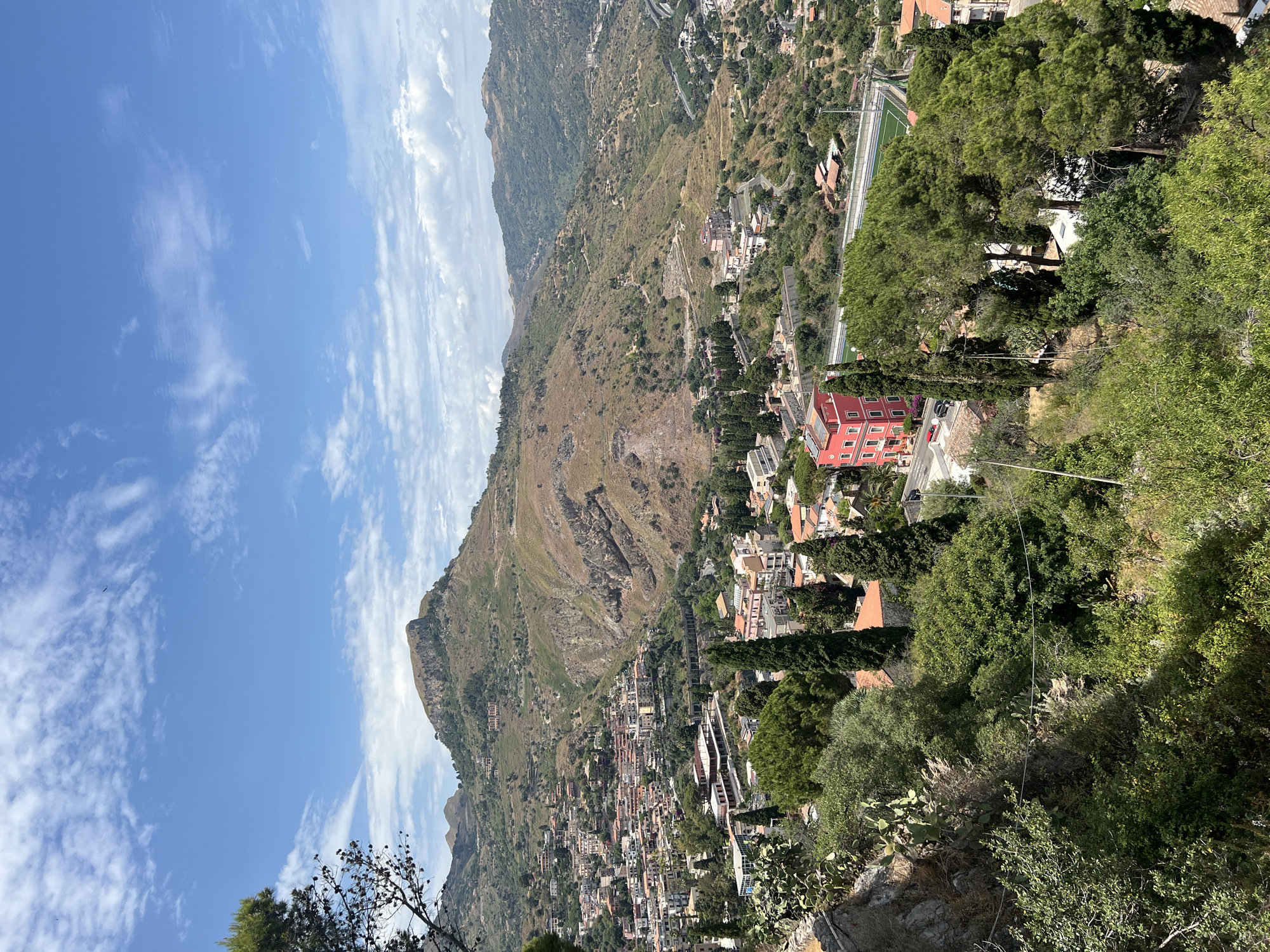 This is a high-angle, full-shot photograph taken on a bright, sunny day in Taormina, Italy, likely from a viewpoint overlooking the town and surrounding hills. The scene is dominated by a sprawling hillside town with buildings of various colors and architectural styles, featuring terracotta roofs. The town is nestled against a backdrop of undulating, arid mountains, with the peaks of the mountains receding into the distance under a blue sky dotted with white, wispy clouds.
In the foreground, a lush green hillside is covered with dense vegetation, including trees, bushes, and some cactus plants growing on a rocky outcrop. A prominent, bright red building with multiple stories stands out amongst the other structures in the middle ground, adjacent to a sports field with artificial turf. Cypress trees are scattered throughout the landscape, adding vertical accents to the scene. Further into the distance, the town stretches along the slopes of the hills, with roads and infrastructure visible. A winding road is visible in the upper right portion of the image, leading towards more distant hills that are covered in green vegetation. The overall impression is one of a vibrant, historic town set within a dramatic natural landscape.