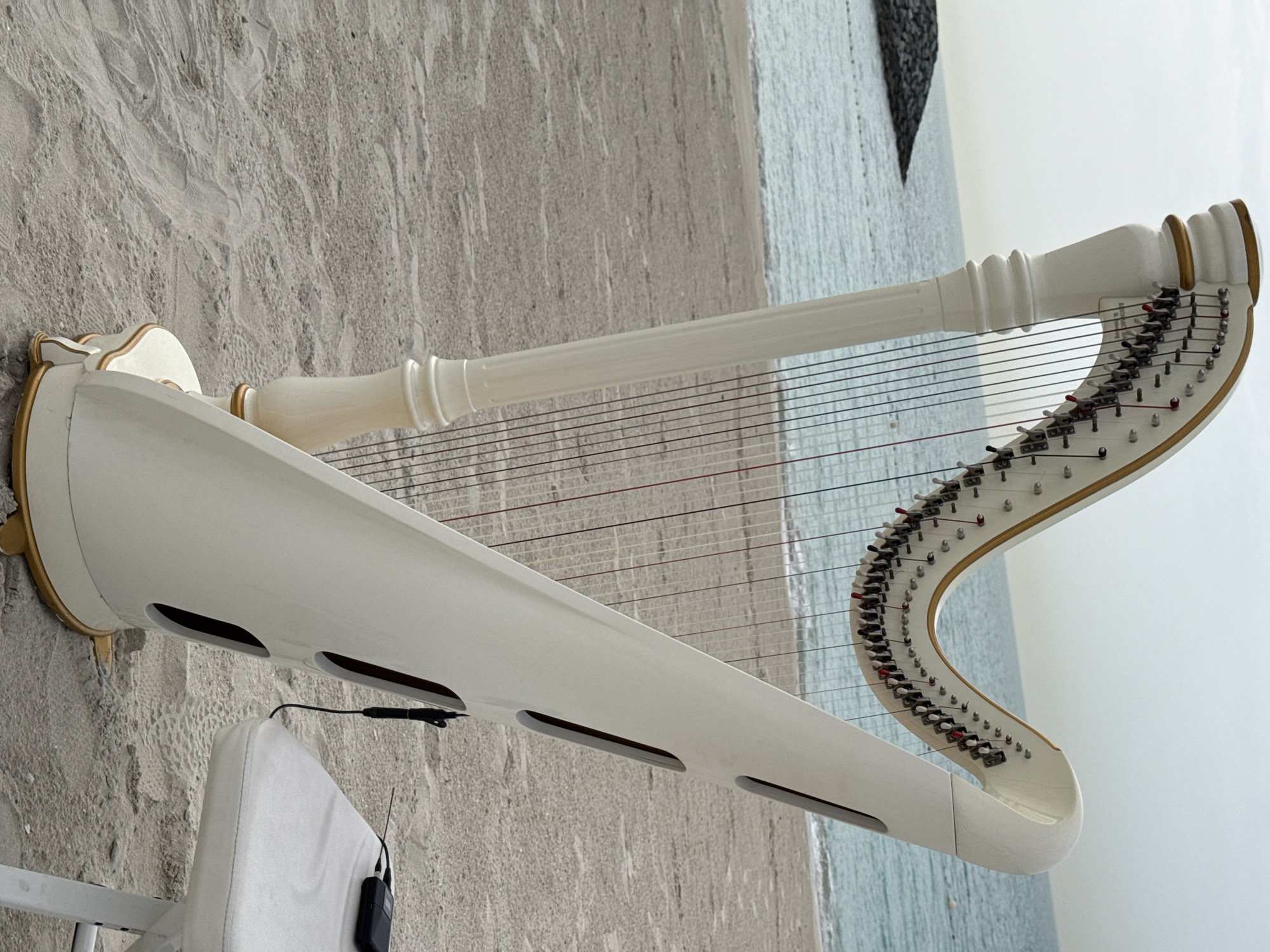 A white and gold concert harp sits on a sandy beach, likely at Ghadeer Al Tayr, United Arab Emirates, based on the provided location. The harp's soundbox is angled towards the lower left of the image. The harp's strings stretch towards the upper right. A white chair and microphone are positioned near the soundbox. The backdrop includes beach sand and the ocean. The sky is overcast.