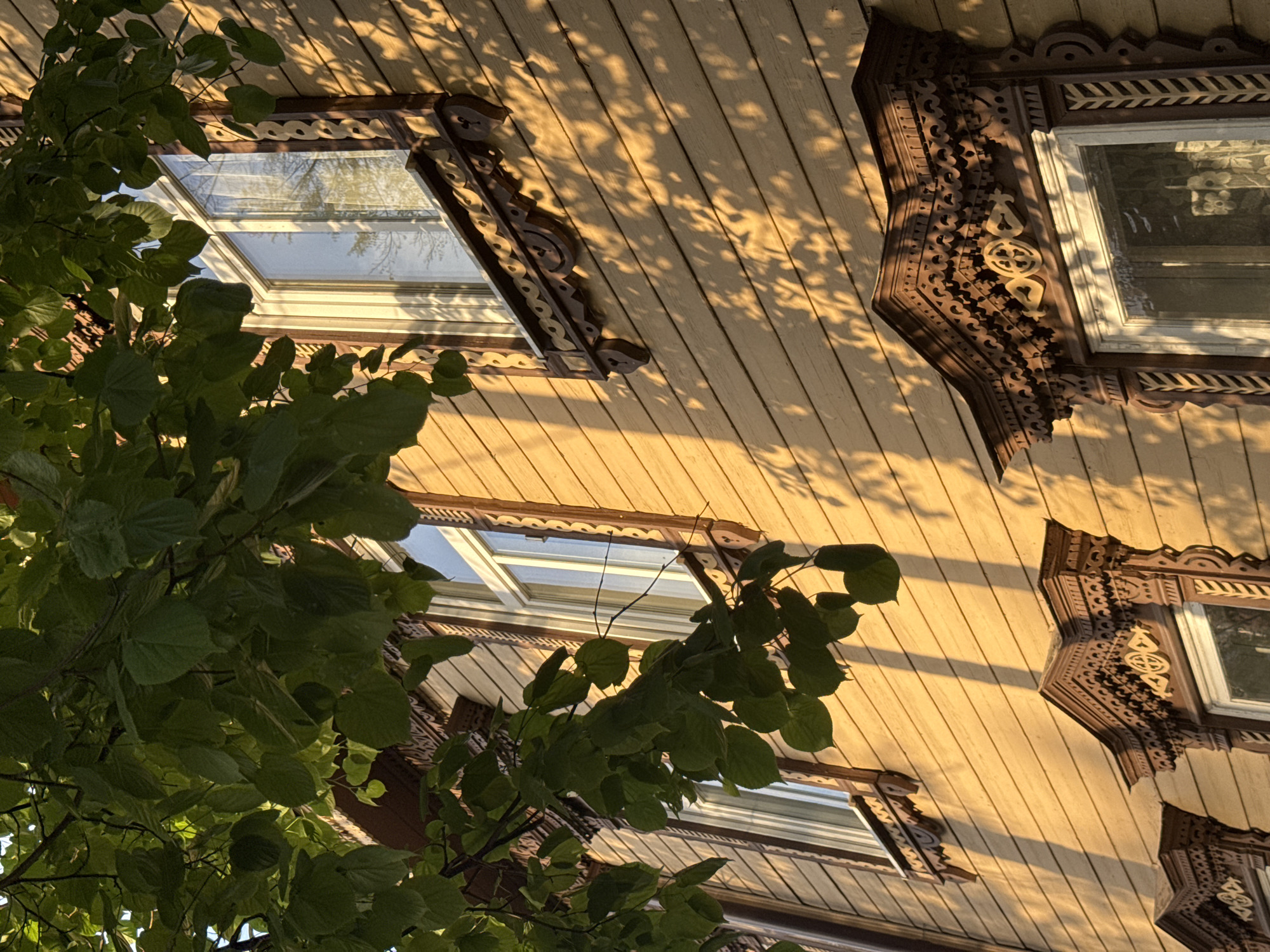 The image shows a low-angle view of a building facade, possibly in Kazan, Russia, with ornate window frames and light-colored wooden plank siding. Three windows are visible, each adorned with detailed, dark-stained wooden carvings. A leafy tree branch partially obscures the lower left portion of the image. The lighting suggests late afternoon or early morning. Shadow patterns from foliage are cast on the siding.