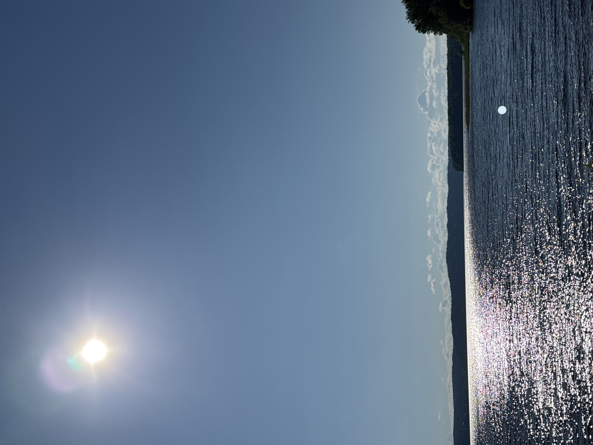 The image depicts a wide, tranquil body of water, likely a lake or large river, under a clear blue sky. The sun, positioned in the upper left quadrant, casts a bright glare and a faint halo effect on the sky. Its rays create a shimmering, sparkling path across the dark blue water, with the light reflecting off countless small ripples and waves. In the distance, a dark, silhouetted treeline with rolling hills forms the horizon. The shoreline on the right side of the frame is marked by dense green foliage, likely trees. There are no visible people or structures in the image. The overall atmosphere is serene and natural, suggesting a peaceful day in a rural or semi-rural outdoor setting. The specific location in Gora (Khokhlovskoe S/P), Russia, is implied by the metadata or context from which the image was provided.