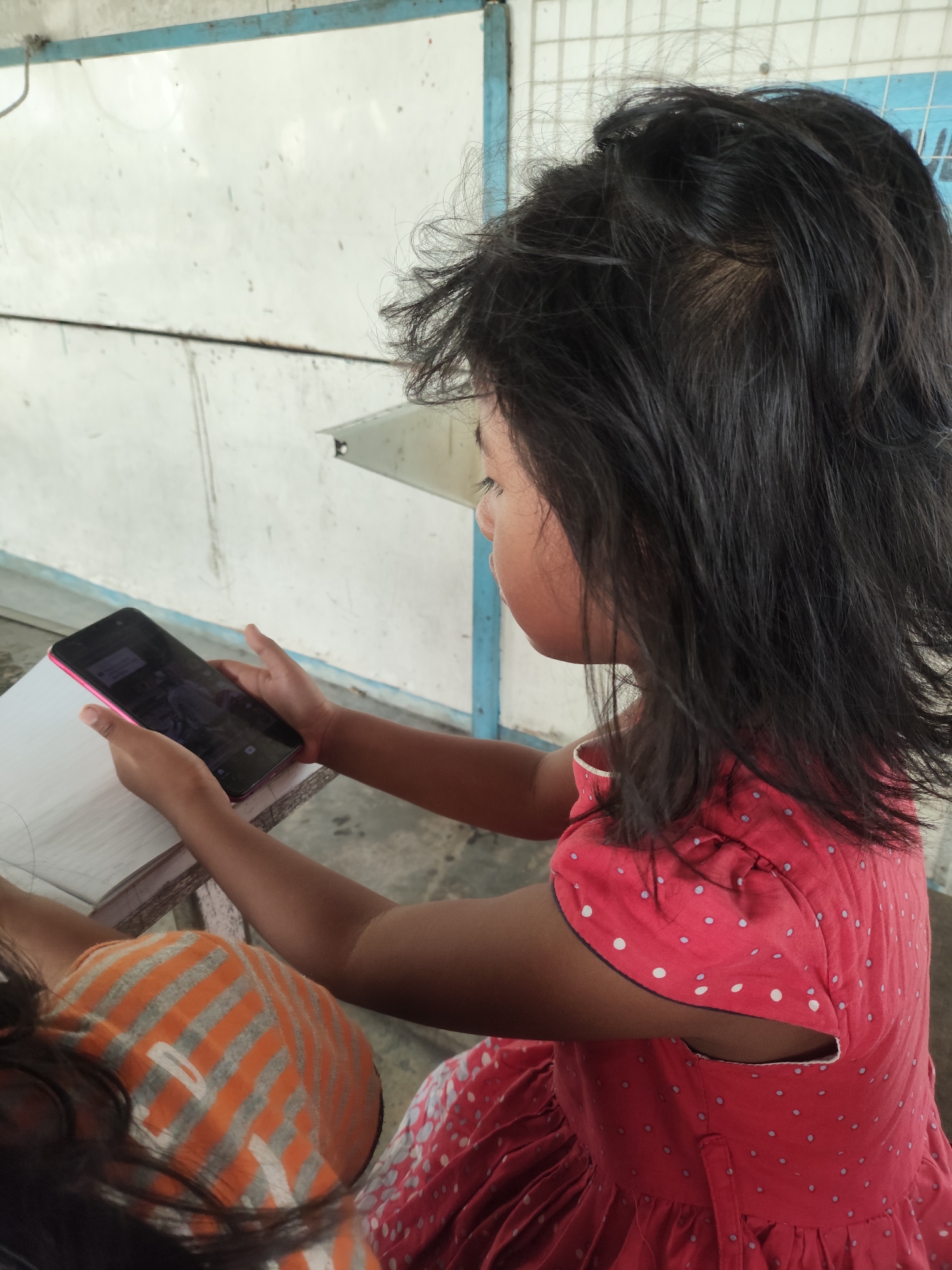A young Indonesian girl, with dark hair and wearing a red polka dot dress, sits at a table and looks at a smartphone. She is in a room with a whiteboard in the background. The lighting is natural and appears to be from overhead. The girl is sitting at a table and holding a smartphone in her hands. A notebook is also on the table. Another person is partially visible in the foreground, wearing an orange and gray striped shirt. The overall scene suggests a casual setting, possibly a classroom or home.