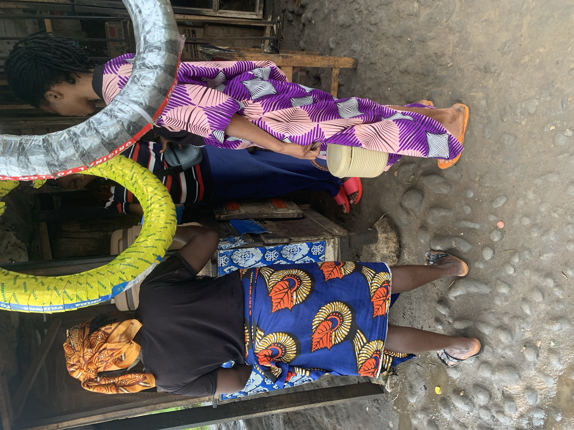 In Shagamu, Nigeria, three individuals are visible outdoors in what appears to be a market or roadside stall. The foreground shows the backs of two women. The woman on the left wears a black t-shirt and a blue patterned wrap skirt with orange and yellow motifs. She has an orange patterned headscarf. The woman on the right is dressed in a purple and pink patterned robe and holds a beige woven basket. Her feet are shod in orange flip-flops. Behind them, another person in a striped shirt and blue trousers stands near a stall exhibiting two large, circular objects wrapped in yellow and grey plastic, possibly tires or similar industrial goods. The ground is uneven and appears wet, with stones and some debris. The overall environment suggests an outdoor commercial setting in a town.