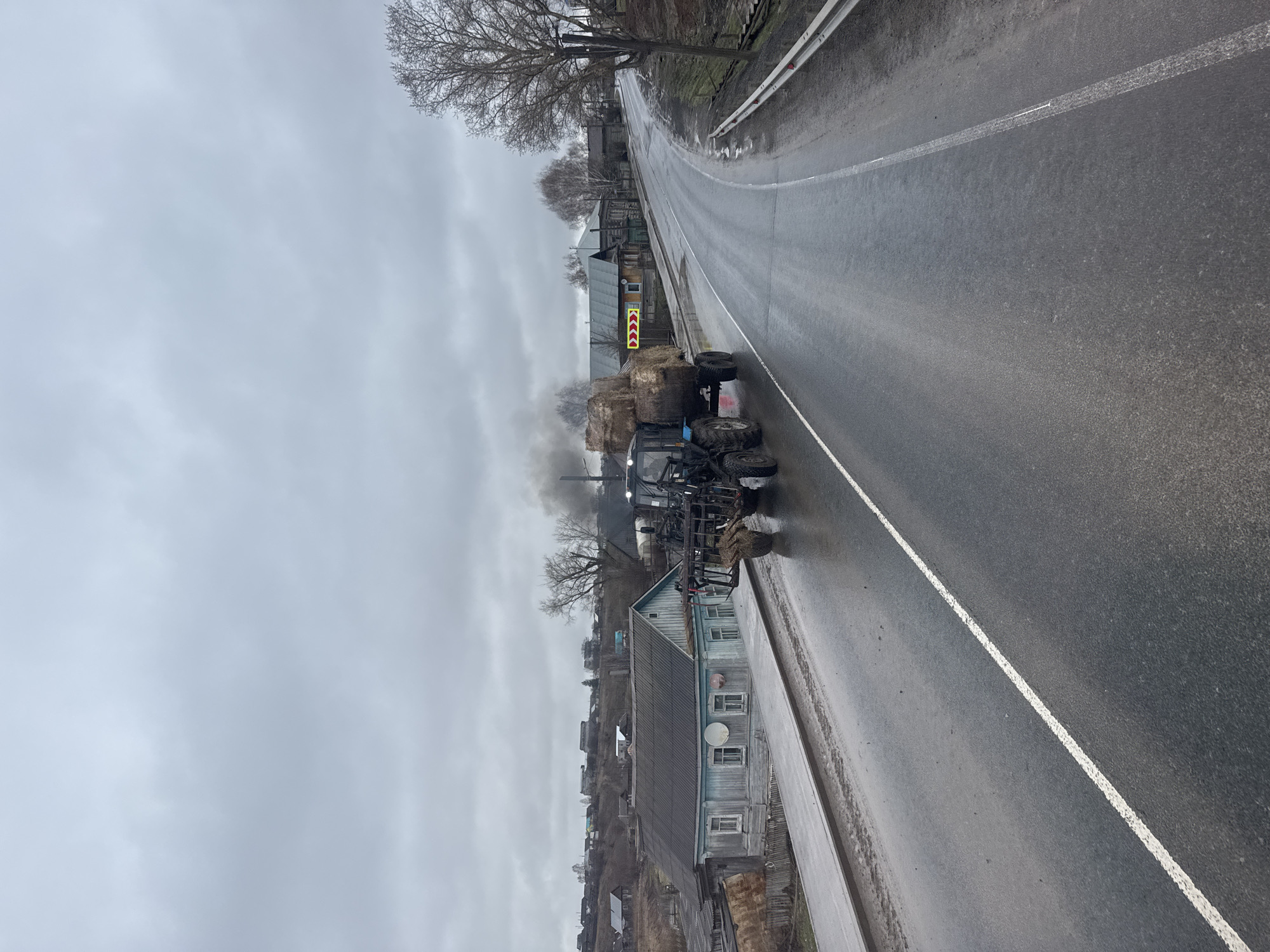 The image captures a rural or semi-rural road scene under an overcast, gray sky, suggesting a cool or cold day. The asphalt road, wet from recent precipitation, stretches into the distance, marked by a continuous white line along its edge.
In the foreground, a blue tractor with a front-end loader is parked or moving slowly on the right side of the road. Dark smoke billows from its exhaust pipe, indicating it is active. Behind the tractor, a trailer loaded with large, rounded hay bales is visible.
Along the left side of the road, a line of traditional wooden houses with pitched roofs can be seen. One prominent house is painted light blue with a darker gray roof and features multiple windows and a satellite dish on its facade. Bare trees, stripped of their leaves, dot the landscape, suggesting late autumn, winter, or early spring. A yellow and red chevron sign, typically indicating a sharp curve or hazard, is visible further down the road, past the tractor. The overall setting points to a small town or village road in Богородск, Russia. No specific people are visible.