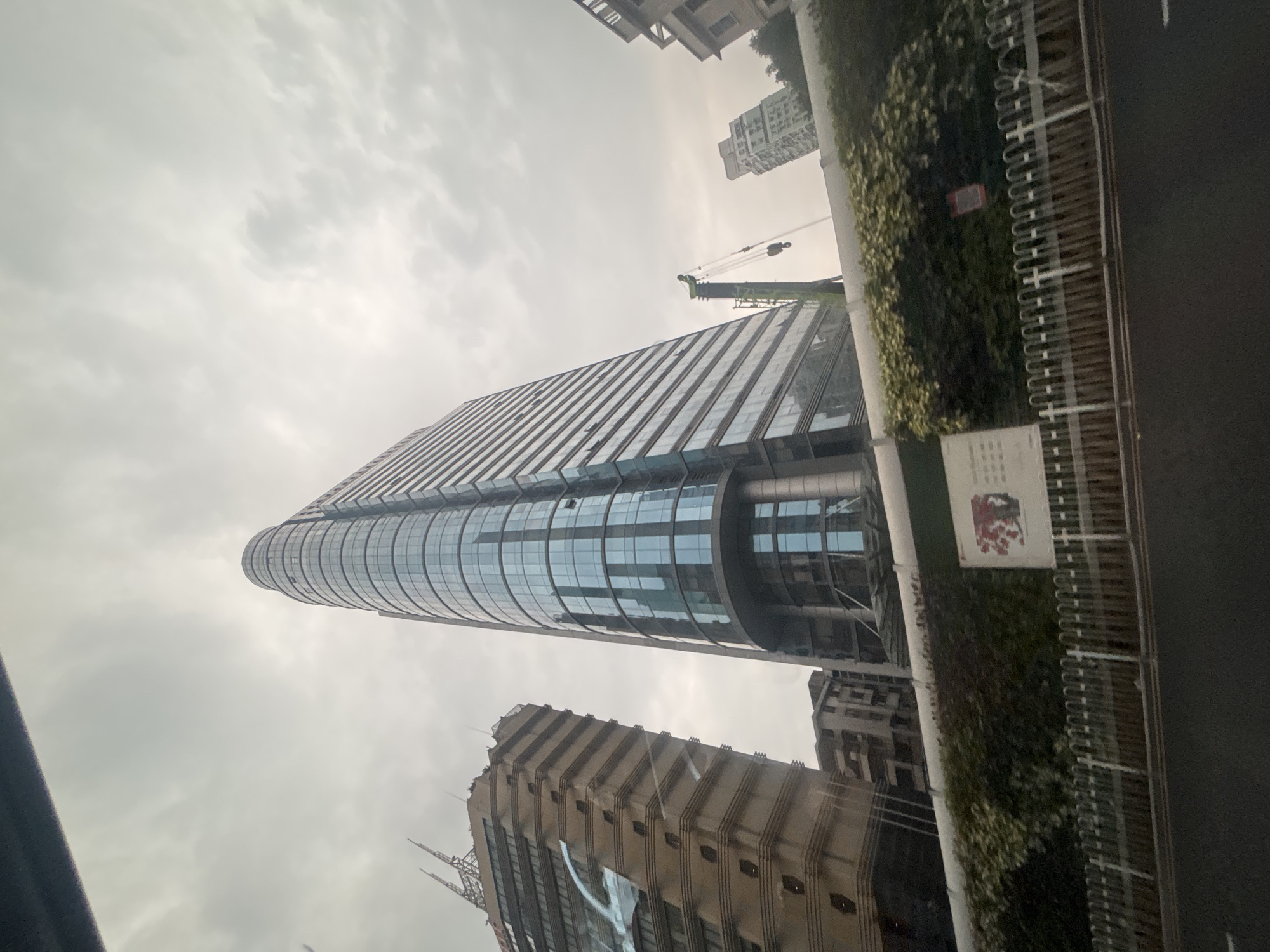 The image captures an urban street scene in Yuexiu District, China, under an overcast sky. Dominating the frame is a tall, modern glass-facade skyscraper with a curved, tapering design. To its left stands another, older, tan-colored building with a more angular structure, featuring several antenna-like protrusions at its apex. In the mid-ground, a green construction crane is visible next to the glass skyscraper, with its boom extended. Further in the background, several other buildings of varying heights and architectural styles are present, suggesting a dense urban environment. A lush green wall of foliage runs horizontally across the lower portion of the image, partially obscuring some of the lower structures and a white fence situated in front of it. A street with visible lane markings is in the foreground, indicating a road. A sign with red and white elements is mounted on the green wall. The overall impression is one of ongoing development and a busy city landscape.