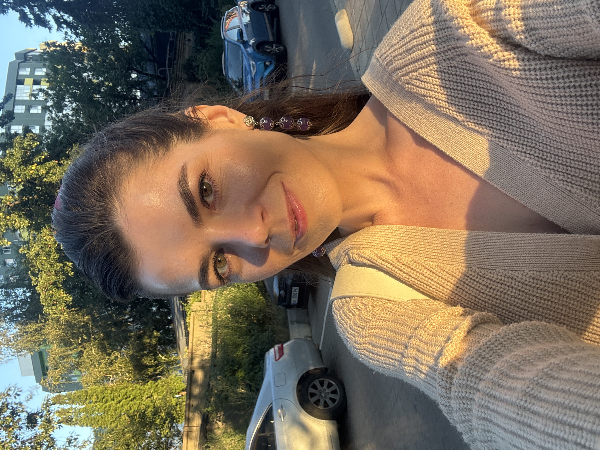 A woman stands in what appears to be a parking lot at dusk, illuminated by the setting sun. She is taking a selfie, her face angled towards the camera. She has brown hair pulled back and is wearing a cream-colored knit sweater. Dangling from her left ear is a long earring with three purple beads. In the background, a silver car is parked to her left, and a blue SUV is parked to her right. Trees and a building are visible behind the cars. The lighting suggests the golden hour, casting warm shadows across the scene. The location is identified as Unknown, Unknown.