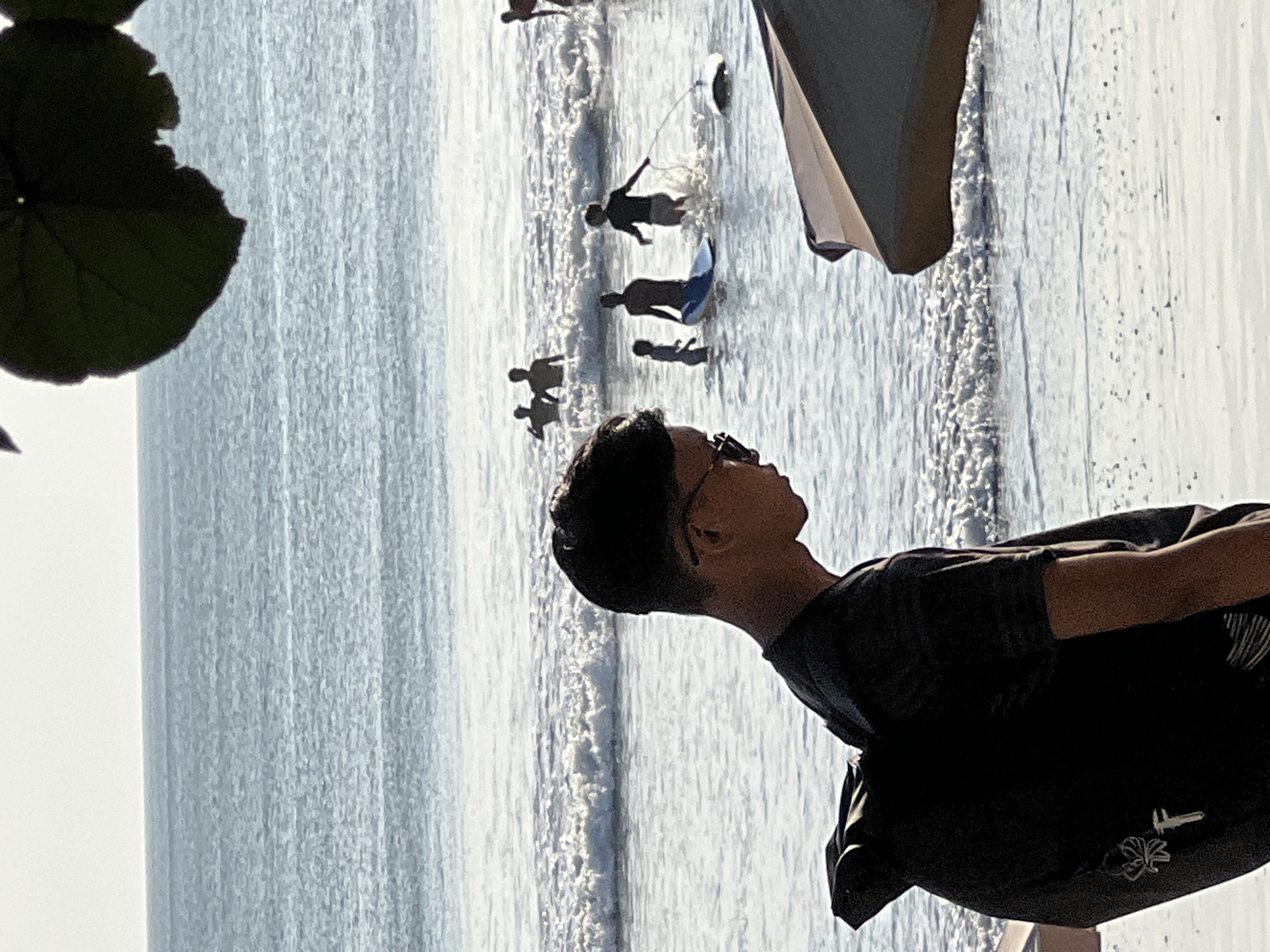 This is a medium shot of a young man wearing a dark t-shirt and sunglasses, with a black backpack on his shoulders, standing on a beach in Kuta, Indonesia. He is facing to the right, with his profile visible. In the background, several people are in the ocean, some are wading in the shallow water and others are in deeper water with surfboards. The waves are breaking on the shore, and the sky is a pale blue. The foreground shows the sandy beach and a portion of an umbrella.
