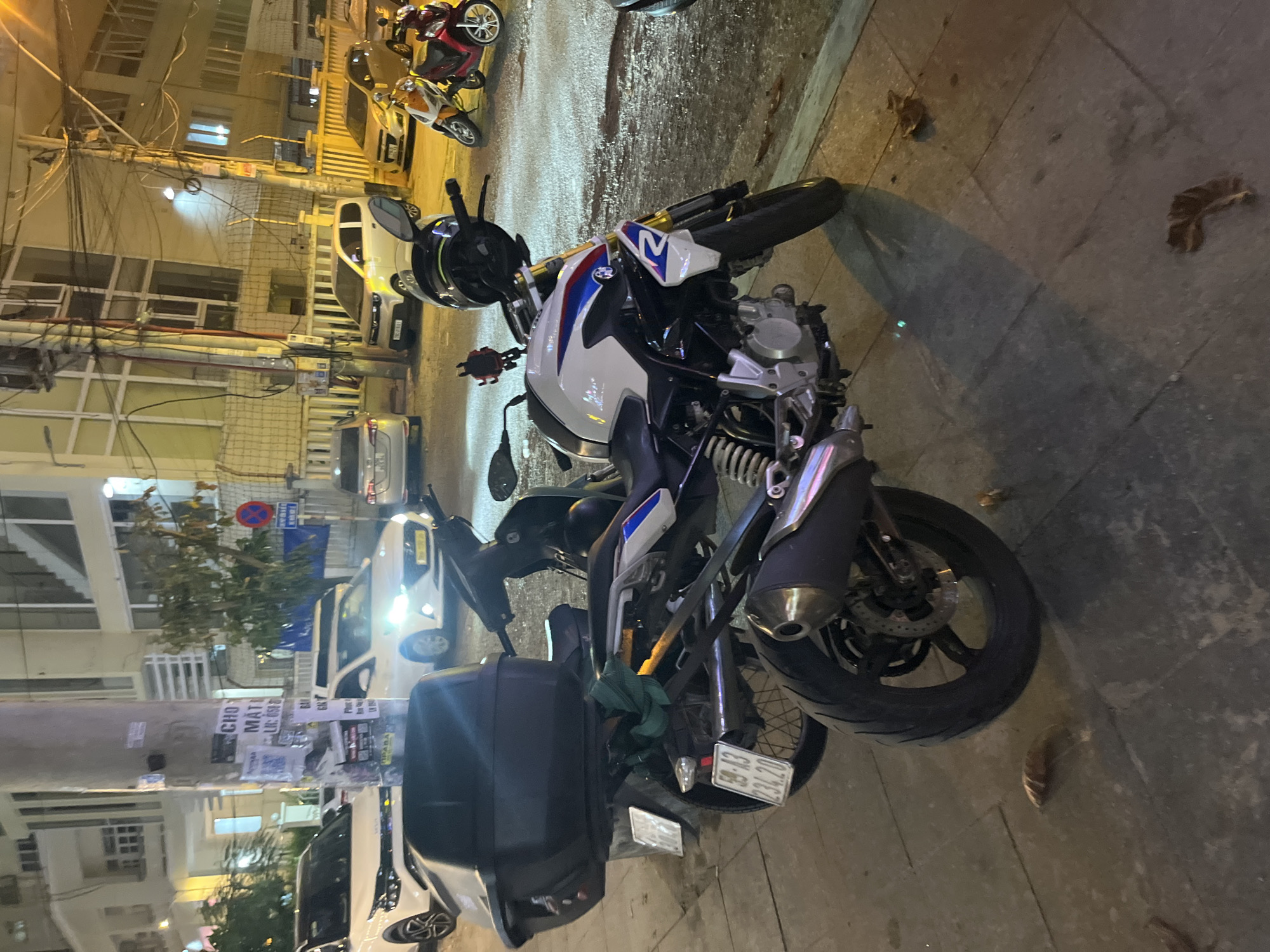 The image depicts a street scene at night in Bac Nha Trang Ward, Vietnam. The foreground is dominated by two motorcycles. The larger motorcycle, a white and blue BMW, is parked on the sidewalk and is the primary focus. It has a black top case mounted on the rear. A black helmet rests on its handlebars. The second motorcycle, a scooter, is partially visible behind the BMW and is black with a green cloth draped over its rear.
Behind the motorcycles, a white car is parked, and further back, other vehicles including a white sedan and two smaller scooters, one orange and white and the other red, are visible on the street. The background consists of buildings, streetlights, power lines, and a sidewalk lined with trees. Some posters are attached to a utility pole on the left side of the image. The ground is paved with concrete slabs. The overall impression is a typical urban street scene at night.