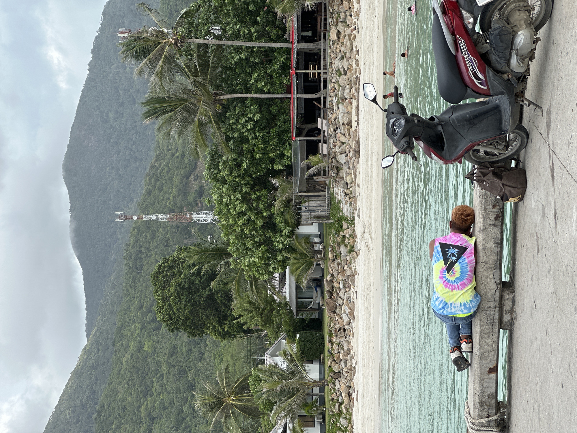 A person in a tie-dye t-shirt and jeans lies asleep on a concrete ledge by the water. A brown backpack rests next to them, and a red scooter is parked nearby. In the background, a sandy beach leads to a line of buildings and lush green mountains under a cloudy sky. Several people are visible in the water, and two cellular towers stand on the mountainside. The scene appears to be located on Koh Phangan, Thailand.
