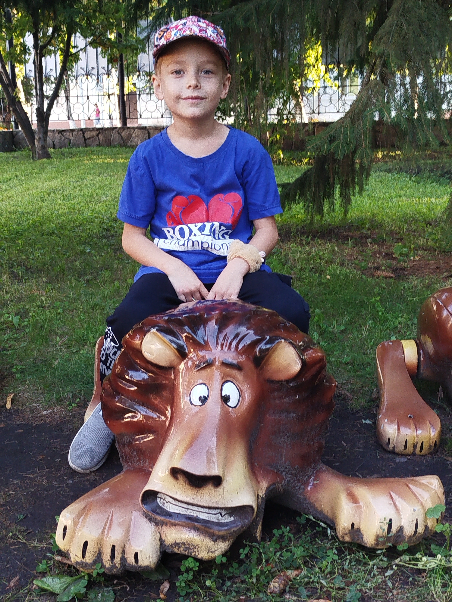 A young boy, approximately 6-8 years old, sits astride a decorative lion statue in a park. He is wearing a blue t-shirt with a "Boxing Champion" graphic, black pants, and a colorful baseball cap. The lion statue is made of brightly colored fiberglass, designed to resemble a cartoonish lion. The boy's hands rest on the statue's mane. His legs are bent, with his feet, wearing gray sneakers, positioned on either side of the statue. The statue is situated on a grassy area with dirt and leaves on the ground. In the background, a metal fence with decorative elements is visible, separating the park from what appears to be a building. Trees with green foliage provide shade. The overall scene suggests a public recreational area within the city of Yalutorovsk, Russia, likely a playground or park designed for children.