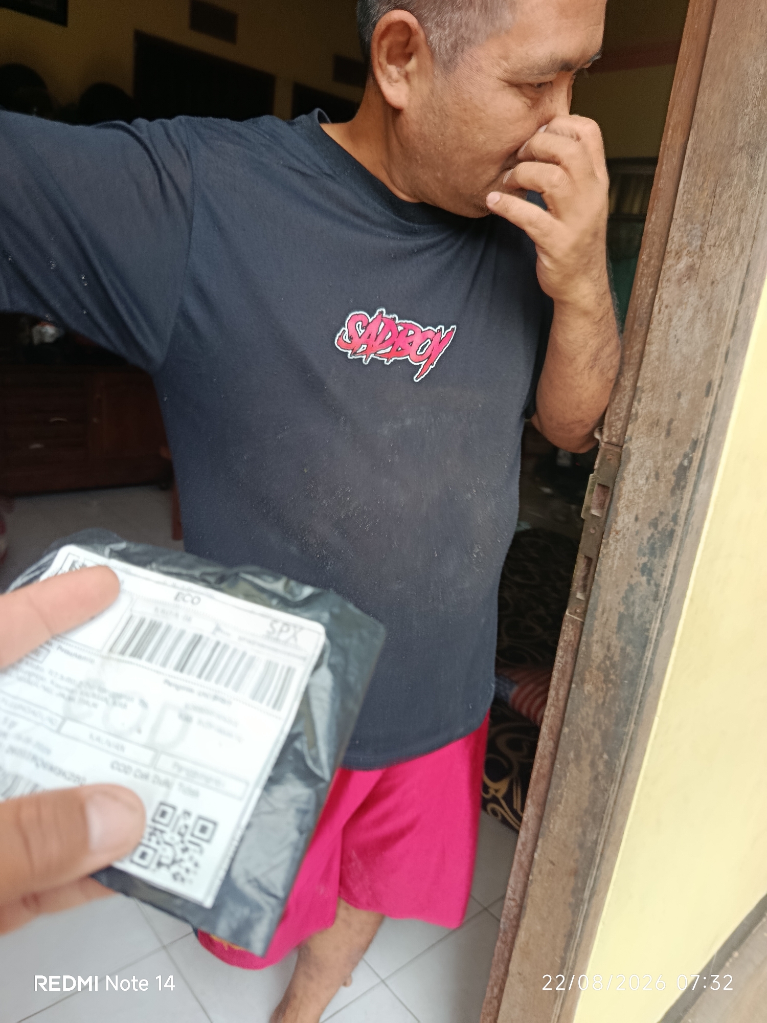 A medium shot captures a man in a black t-shirt with a red "SADBOY" graphic and pink shorts. He is standing in a doorway, holding a small package with a shipping label. The man is touching his nose with his right hand, and his gaze is directed downwards. The environment appears to be indoors, with wooden doorframes and a tiled floor visible. The timestamp "22/08/2026 07:32" is present in the lower right corner. The overall impression is of a casual moment, possibly receiving a delivery.