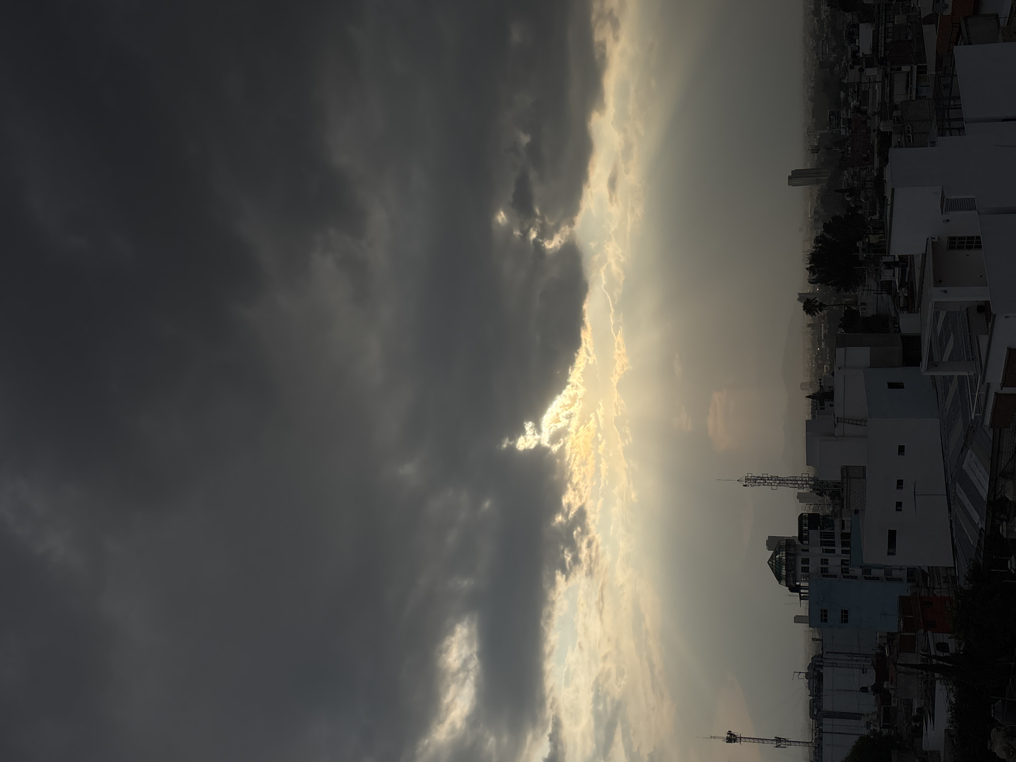 The image shows a view in Puebla, Mexico, at dusk. The left side of the frame is dominated by heavy, dark storm clouds. The clouds on the right side are thinner and lighter, with sunlight filtering through. The cityscape is visible below the clouds. Predominantly white buildings of varying heights are evident, with some taller structures appearing in the distance, including what appear to be cell towers and antennas atop some buildings.