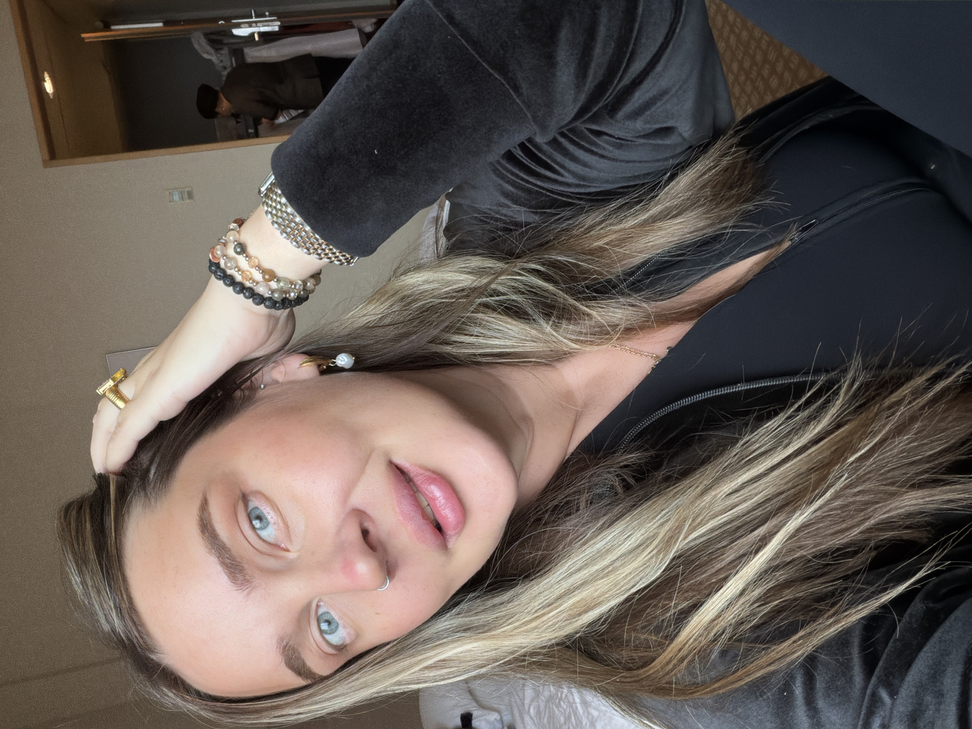 A woman with blue eyes, light brown hair with blonde highlights, and a nose ring is looking at the camera. She is wearing bracelets and a ring on her left hand. She is wearing a black zip-up jacket and a black velvet top. The background is a room with beige walls and a mirror reflecting a person standing. The location cannot be determined to be in Moscow, Russia, based on the visual information provided.