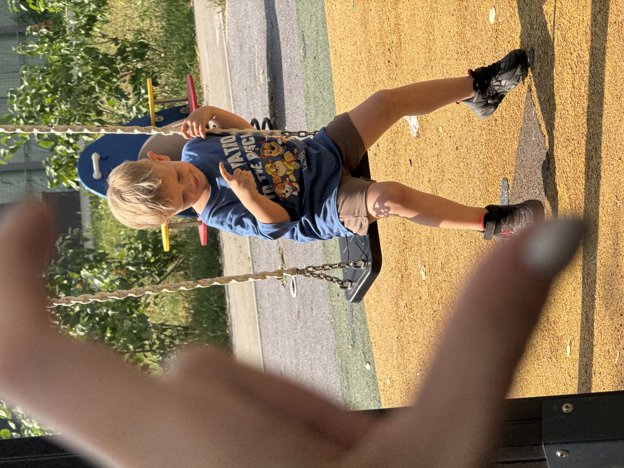 A young boy with blond hair is sitting on a swing at a playground. He is wearing a blue t-shirt with a Paw Patrol graphic and brown shorts. The boy is looking down and pointing his left index finger. He is wearing black sneakers with red accents. The swing has a black seat and is suspended by thick ropes and metal chains. The playground surface is a textured yellow material. In the background, there is playground equipment, including a blue and red structure, and green foliage. The scene is outdoors and appears to be a sunny day. The foreground shows a blurred hand or arm obscuring part of the left side of the image. The location is identified as a playground in Moscow, Russia.