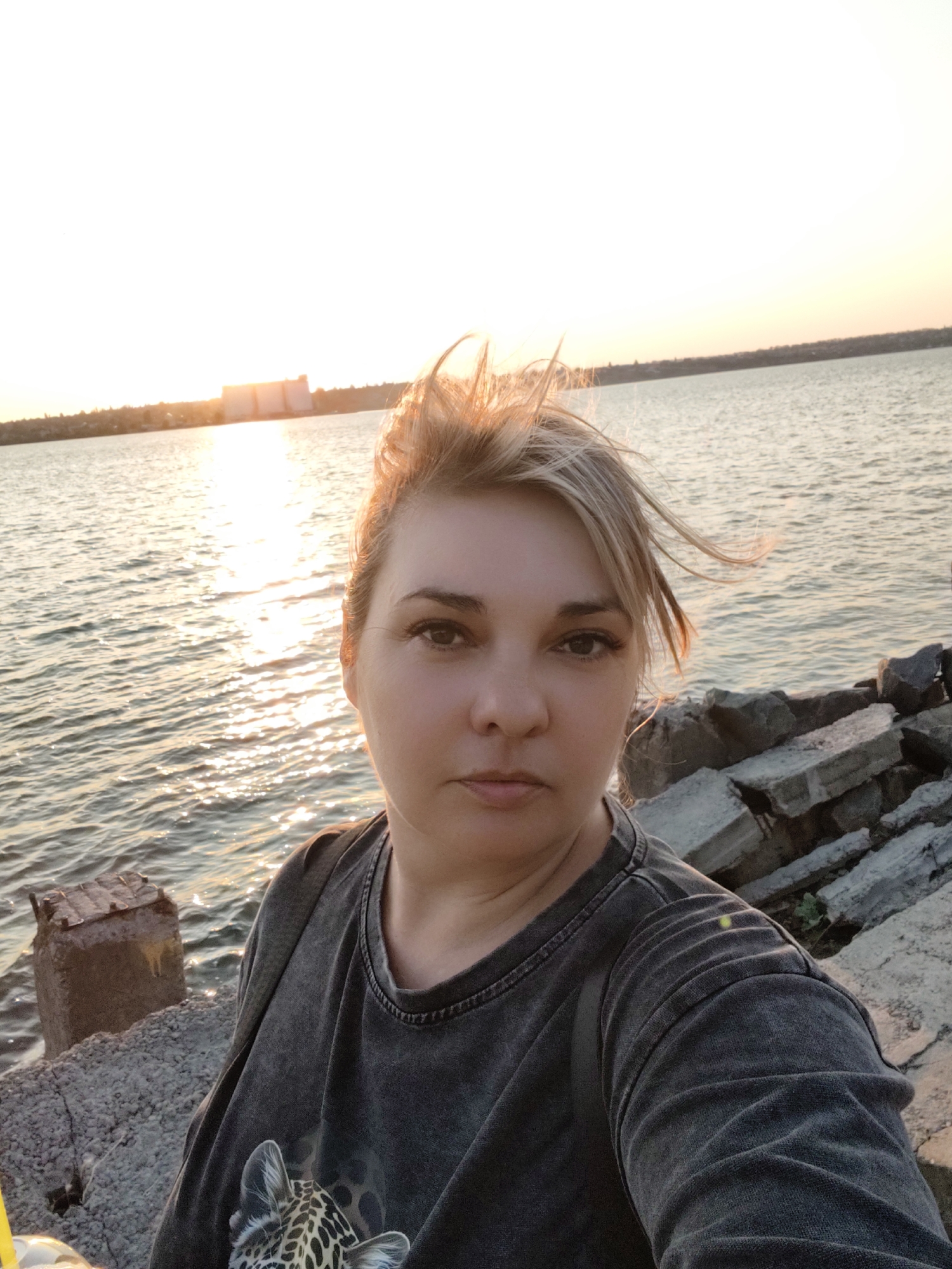 This is a portrait of a woman taken in Mykolaiv, Ukraine. She is standing on a rocky embankment near a body of water, likely a river or the sea, with a large building visible in the distance on the opposite bank. The sun is setting, casting a warm golden light across the water and the sky, creating a peaceful and serene atmosphere. The woman is looking directly at the camera with a neutral expression. Her blonde hair is windswept, suggesting a gentle breeze. She is wearing a dark grey t-shirt with a leopard print detail on the chest. The foreground shows some of the rocky terrain and a concrete structure. The image captures a quiet moment by the water at dusk.