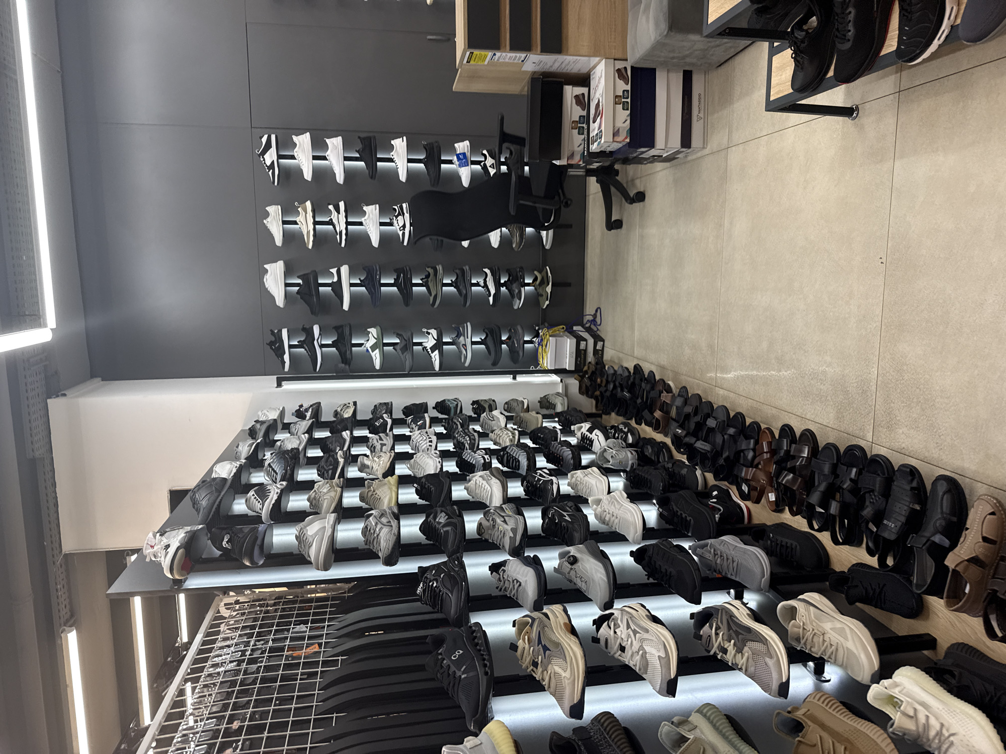 The image depicts the interior of a modern shoe store. The scene is dominated by rows of display shelves showcasing a large variety of athletic footwear, primarily sneakers. The shelves are organized in a grid-like fashion, with shoes neatly arranged on each level. The lighting in the store is bright, emanating from overhead fluorescent fixtures.
On the left side of the image, a metal rack is visible, possibly for hanging clothing or accessories. Further back, a section of the store appears to be dedicated to bags or other retail items.
In the center of the image, a black ergonomic office chair is positioned in front of a wall of shoe displays. The wall is a dark grey, creating a contrast with the lighter colored shoes. There are stacks of shoe boxes on the floor to the right of the chair, some of which are open, revealing shoe boxes with branding such as "MARCOshoes".
The flooring is a light-colored tile. The overall impression is one of a well-stocked and organized retail environment, likely a shoe store catering to a wide range of styles and brands. The image does not contain any people.
This image was taken in Chișinău, Moldova. The specific store is likely to be "MARCOshoes", a prominent shoe retailer in Moldova, indicated by the presence of their branded boxes.