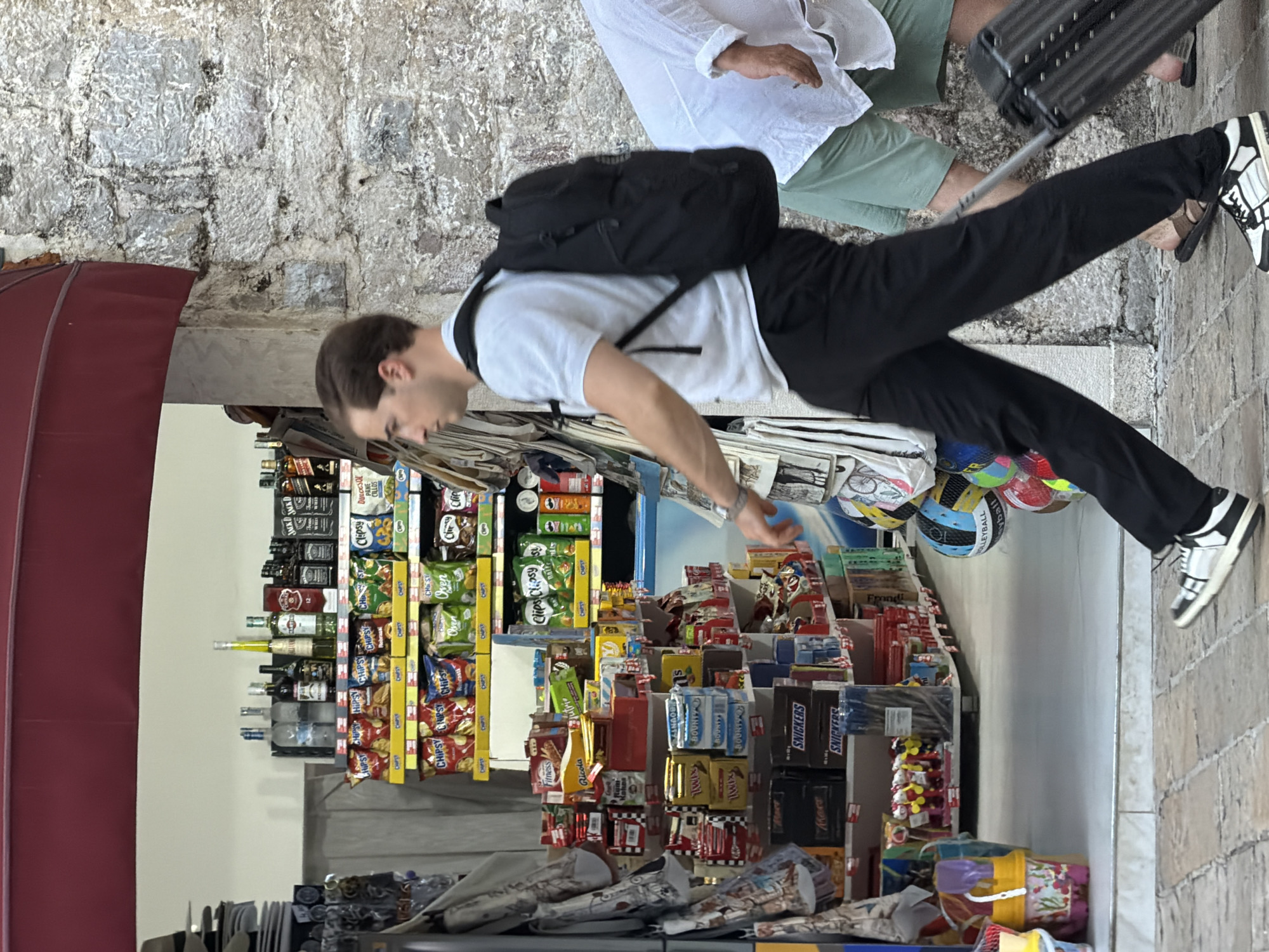 A young Caucasian man with short brown hair is walking in front of a store in Budva, Montenegro. He is wearing a white t-shirt, black pants, and white sneakers with black laces. He has a black backpack on his back and is pulling a black suitcase with his right hand. The store appears to be selling snacks, drinks, and souvenirs. There are shelves stocked with various products, including chips, candy bars, and bottles of alcohol. In the background, another person is visible wearing a white shirt and green shorts. The man is walking on a cobblestone sidewalk. The store has a red awning and a stone facade.