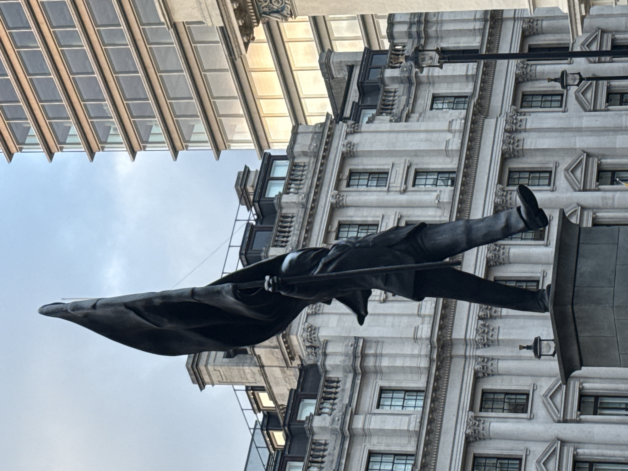 The image shows a bronze statue of Colin Campbell, 1st Baron Clyde, in Waterloo Place, London, United Kingdom. The statue depicts Campbell standing in military uniform, holding a flag. Behind the statue are two buildings. One building is modern with repetitive rectangular windows. The second building is older and more ornate, with classical architectural details and rows of windows. The sky is visible in the background and is mostly overcast.