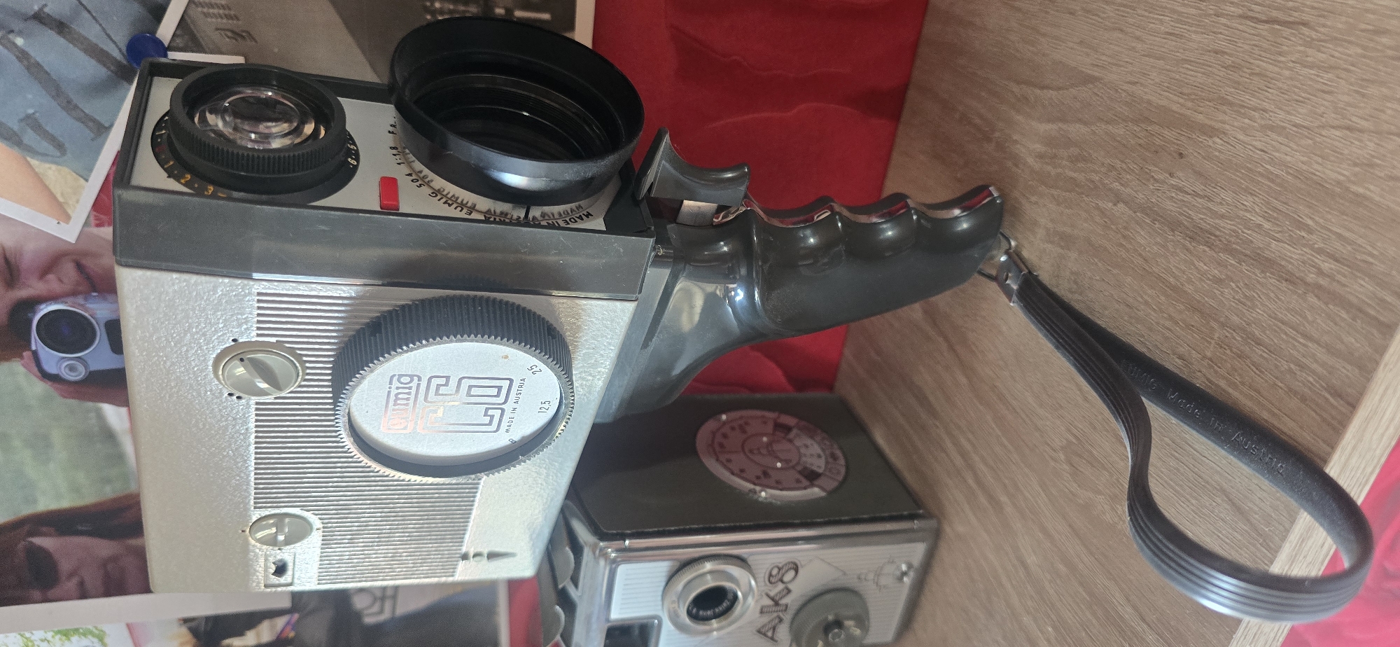 The image displays a collection of vintage cameras, primarily a silver and black Eumig film camera with a prominent lens and handle. To the left, a smaller, silver camera with "AK8" inscribed on its side is visible. The cameras are arranged on a wooden surface, with a red backdrop behind them. The Eumig camera has a ribbed grip and a strap attached to its handle. The overall scene suggests a display or collection of photographic equipment. There is no indication of the city of Gostagayevskaya, Russia, in the image.