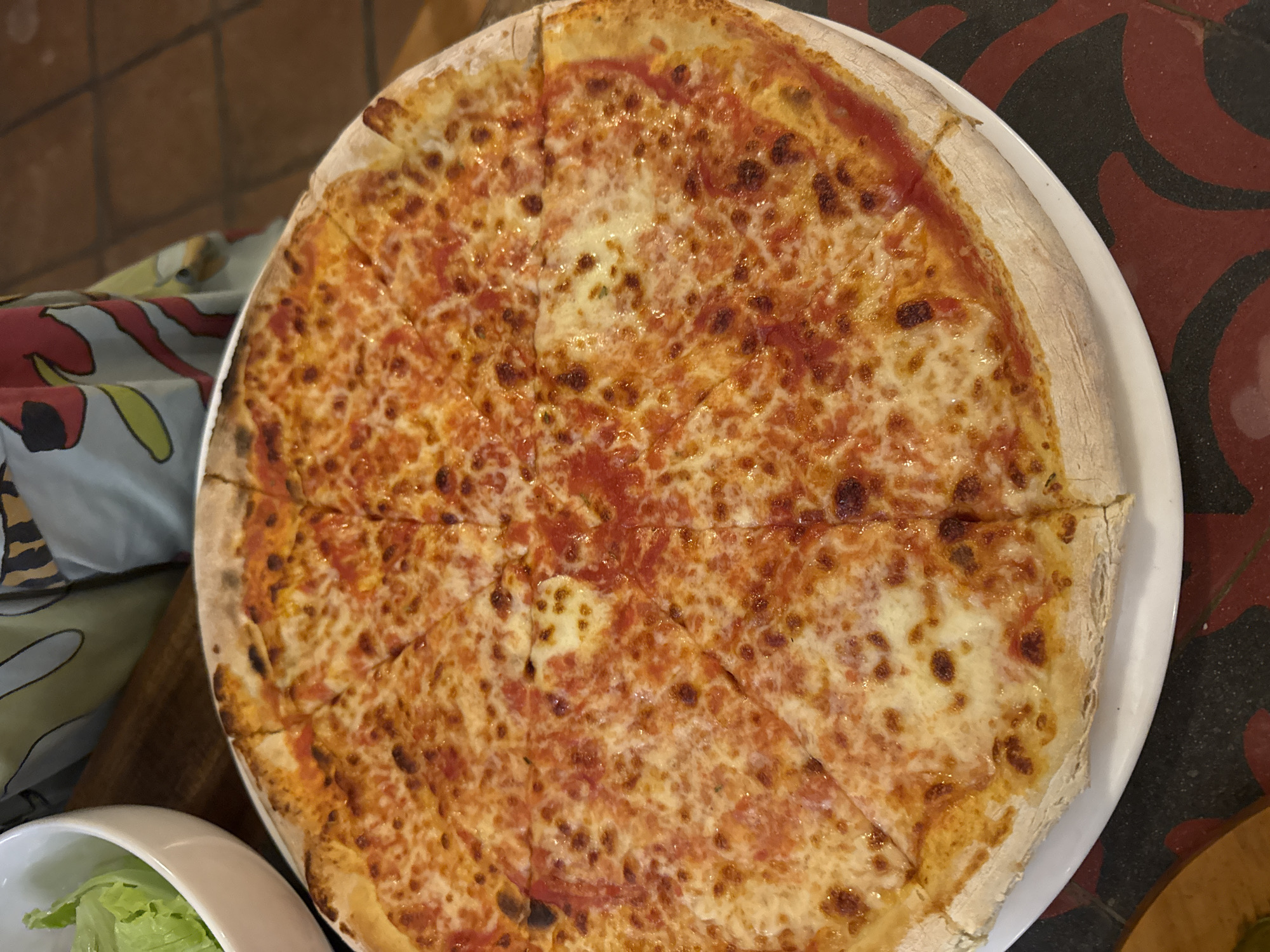 The image shows a circular pizza on a white plate. The pizza is cut into several slices and appears to be a Margherita style, with tomato sauce and melted cheese. The crust is visible, and the pizza shows some browning from baking. The plate is placed on a table with a patterned tablecloth or placemat nearby, as well as another bowl containing green lettuce. This picture was taken in the city of Ungasan, Indonesia.
