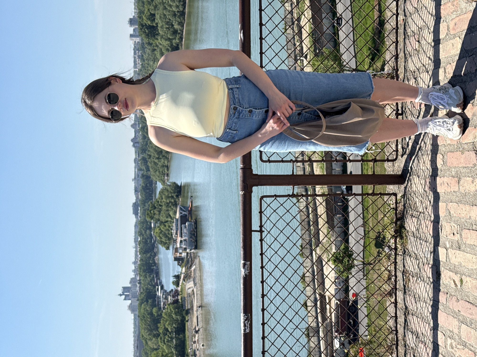 A woman wearing sunglasses, a yellow top, and denim shorts stands on a red brick structure overlooking a river. She holds a brown bag. The background includes a cityscape, trees, and boats on the river. A metal railing runs along the structure. The location is likely in Belgrade, Serbia, judging by the architectural style and urban environment.