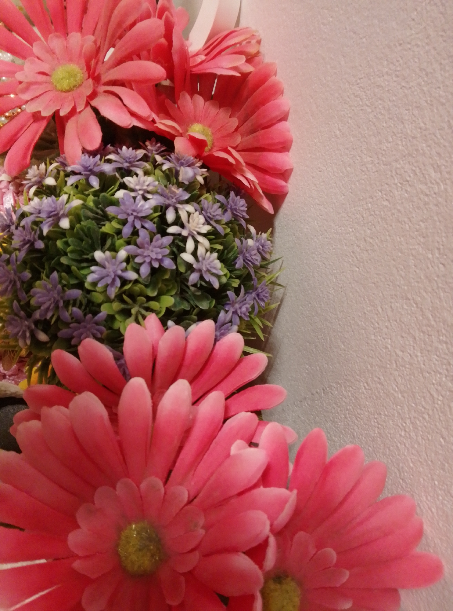 This is a close-up, eye-level shot of artificial flowers against a textured, light-colored wall. The arrangement consists of large, pink daisy-like flowers and a rounded cluster of smaller, purple and white flowers with green foliage. The flowers are the primary subjects, filling the left and lower portions of the frame. The background wall occupies the right and upper portions of the image. No people are present. There is no specific information to ascertain the location within the city of Wedel, Germany.