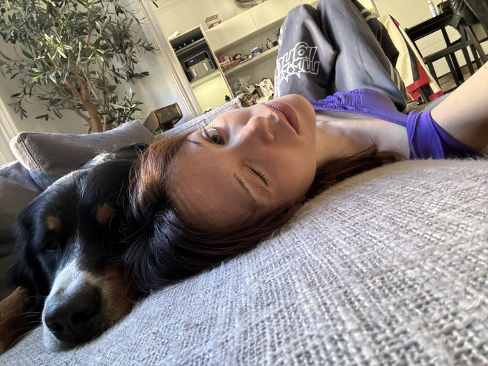 The image displays an interior scene, presumed to be located in Barcelona, Spain. A female individual and a tricolored dog are resting on a light gray, textured couch. The female, with brown hair, wears a purple top with ruched detailing and gray sweatpants featuring white lettering on the left leg; she is positioned on her back, head turned towards the viewer, with her right eye open and left eye partially closed. The dog, marked with black, white, and tan fur, lies adjacent to the female on the couch, its head resting near her upper body, eyes appearing closed. In the background, a large potted olive tree stands on the left. To the right, a white open shelving unit displays various items, including a metallic kitchen appliance, and a dark chair is partially visible on the far right. The environment is brightly illuminated.