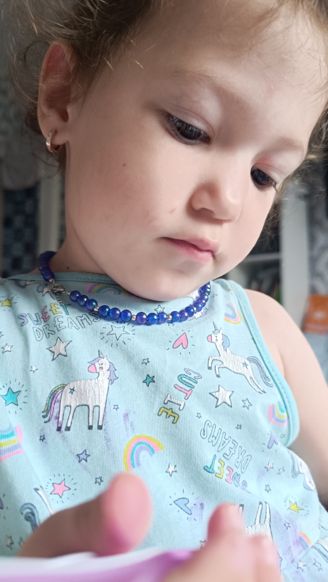 A close-up, eye-level shot focuses on a young child's face and chest, angled downwards as if looking at something in their hands, which are blurred in the foreground. The child has light skin and dark eyes, with a small gold hoop earring visible in their left ear. They are wearing a light blue sleeveless top adorned with a repeating pattern of unicorns, rainbows, stars, and text that appears to read "SWEET DREAMS" and "CUTE". A necklace made of small, iridescent blue beads is visible around the child's neck, featuring a small silver charm that resembles a star with a chain. The background is out of focus, with hints of patterned wallpaper and shelving with books. The lighting is soft, suggesting an indoor setting. No specific location in Chaikovsky, Russia, can be determined from the visual information provided.