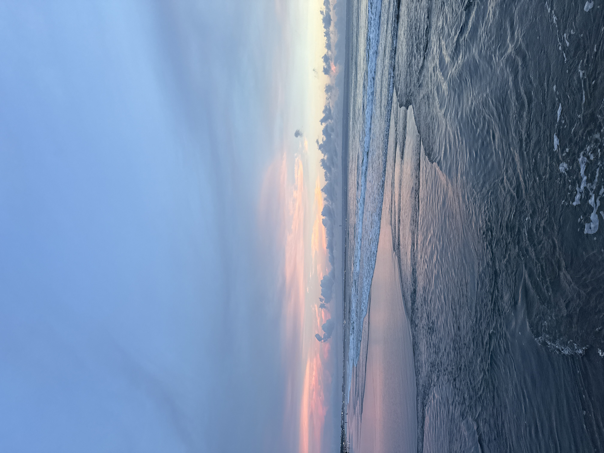 The image depicts a view of Kuta Beach, Kuta, Bali, Indonesia at twilight, looking from the beach towards the sea. The sky exhibits blue, gray, pink, and gold cloud formations illuminated by the setting sun. The shoreline consists of sand meeting the ocean water, which exhibits wave activity with visible foam. The water color varies from light to dark blue. The image is oriented vertically, with the sky dominating the left side and the ocean on the right. Some city lights are visible along the distant horizon.