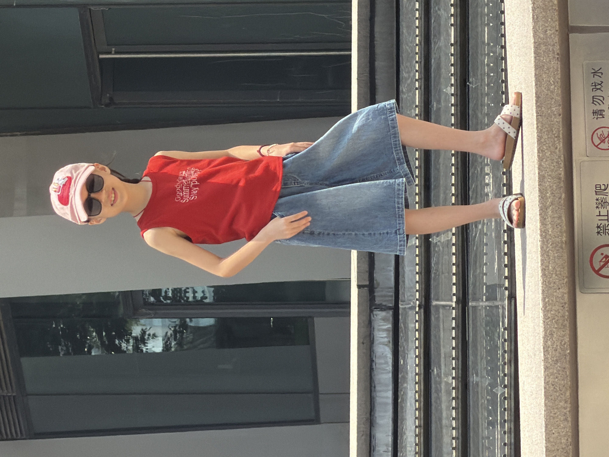 A young woman, facing the camera, stands in what appears to be an outdoor urban setting. She is wearing a pink baseball cap with a graphic on the front, dark sunglasses, a red sleeveless t-shirt with white text, and a light-wash denim skirt. Her attire suggests a casual, warm-weather outfit. She is also wearing white sandals with a patterned strap.
The woman is positioned in front of a modern building with large, dark-tinted windows. To her right, there is a textured concrete wall and a metal structure with vertical slats, possibly part of a railing or a decorative element. A sign with Chinese text is visible on the concrete wall. The text includes characters that translate to "No Playing" and "No Climbing." The image appears to be taken during the daytime, with sunlight casting shadows. The overall atmosphere is bright and relaxed. The woman is smiling slightly, conveying a sense of ease. The Yuzhong District location context places this scene in a busy urban environment in China.