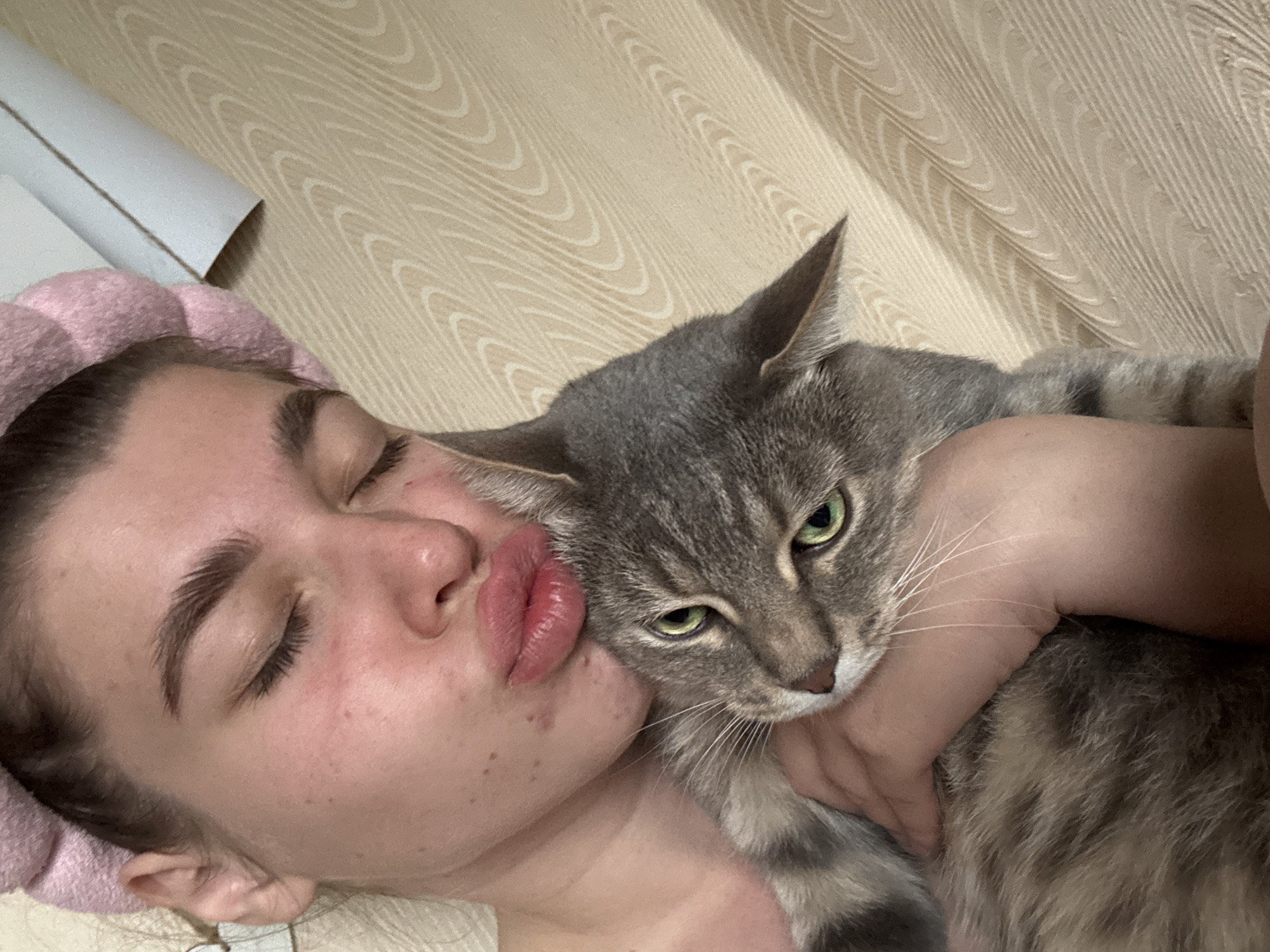 A woman wearing a pink headband lies with her eyes closed, puckering her lips to kiss a gray tabby cat. The cat looks directly at the camera. The scene is indoors, with a patterned wall in the background. The location information is not discernible from the image.