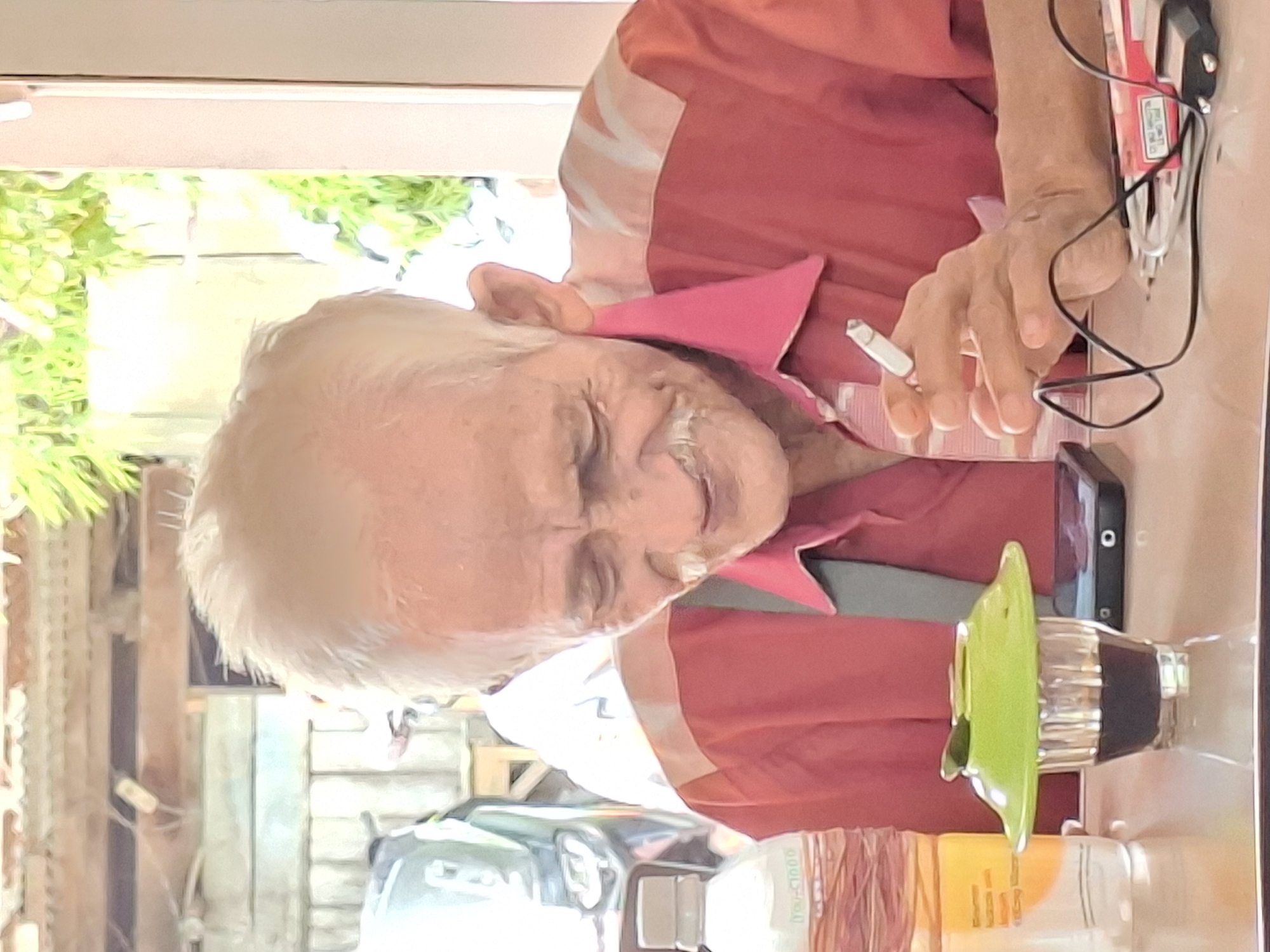 An elderly person with white hair is seated at a table holding a lit cigarette. The individual is wearing a pink collared shirt with white accents. Resting on the table are a smartphone, a bottle with a green top, and medication. In the background, there is a glimpse of a vehicle and trees, likely indicating an outdoor setting. The location may be Lhokseumawe, Indonesia.