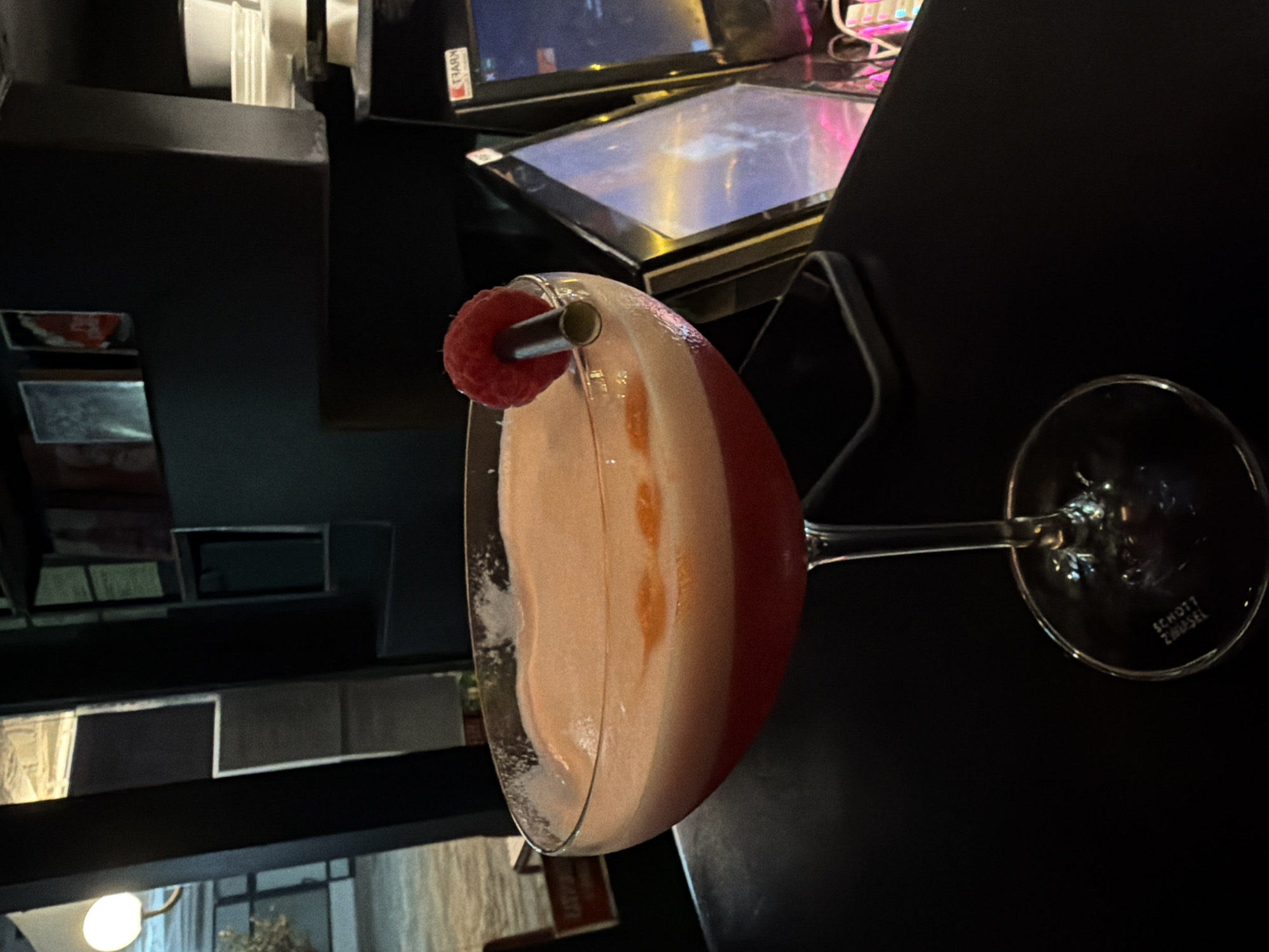 A close-up, eye-level shot features a coupe glass filled with a pink cocktail, garnished with a raspberry and a metal straw. The cocktail has a foamy, light pink top layer and a deeper red bottom layer, with two small orange slices visible in the foam. The glass is set on a dark, reflective surface, likely a table. In the background, a dimly lit environment includes shelves with various items, a wall with framed pictures, and illuminated screens. The lighting is moody, with a warm glow from a nearby lamp on the left. The scene is devoid of people and indicates a bar or restaurant setting in Budva, Montenegro.
