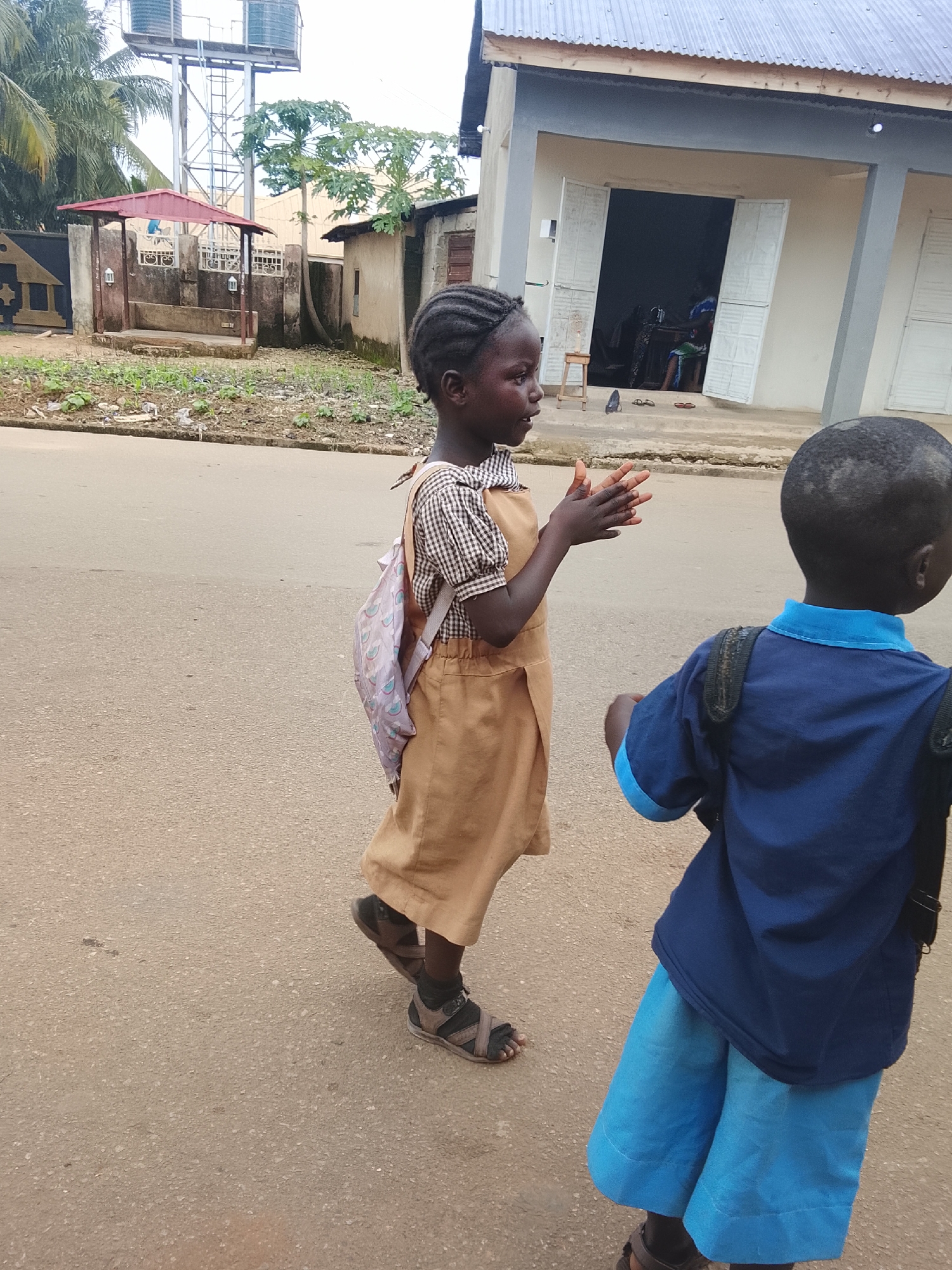Two young children, one in a tan dress and plaid shirt with a patterned backpack, and another in a blue shirt and shorts with a black backpack, are walking on a paved road in what appears to be an urban setting. The child in the tan dress is facing away from the camera and is clapping their hands, while the child in the blue shirt is facing away from the camera. In the background, there is a building with an open doorway, a smaller structure with a red roof, and some vegetation. The overall environment suggests a warm, possibly tropical climate. The presence of the children in school uniforms and backpacks hints at them possibly being on their way to or from school. The scene is captured from a slightly low angle, suggesting the photographer was at a similar eye level to the children.