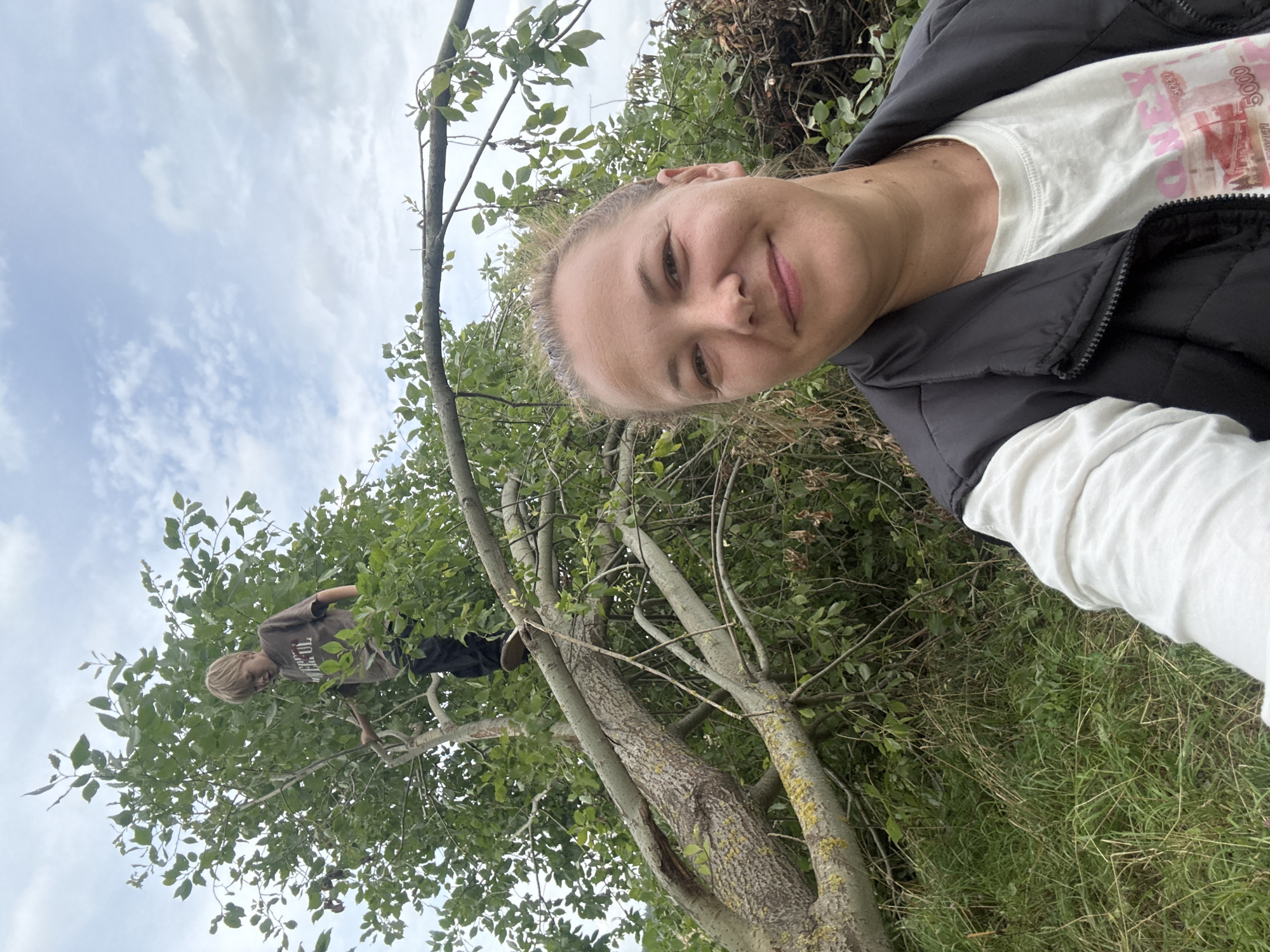 An adult woman with blonde hair pulled back and wearing a black vest over a white t-shirt takes a selfie in the foreground, smiling. Behind her, a young boy with blonde hair is climbing in a large, leafy tree that appears to have a fallen branch across it. The setting is outdoors, with green grass and foliage visible. The sky is overcast with scattered clouds. The image was taken in Dmitrov, Russia.